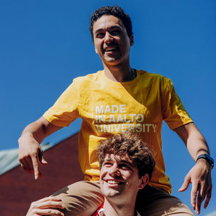 A smiling Aalto student in a yellow Aalto University t-shirt sitting on the shoulders of another student.