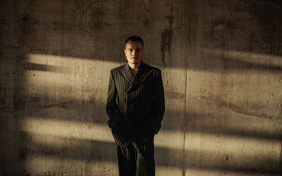 A man in a suit stands in front of a concrete wall with his hands in his pockets