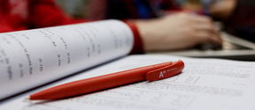 An Aalto pen lying on the page of a study book, students working in the background / photo by Aalto University, Aino Huovio