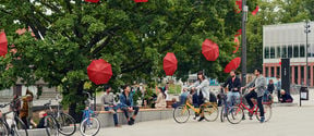 Students on Otaniemi campus. Open red umbrellas attached to trees, students sitting on a bench under the green-leaved trees. Photo by Aalto University /Unto Rautio