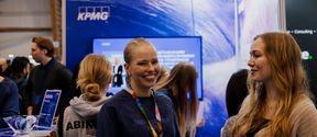 Two women smiling and standing in a crowd. Behing a blue colorful wall with TV screen and KPMG logo on it.