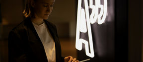 A student scrolling an ipad by an Aalto University logo neon sign.