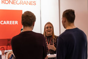 A woman is facing the camera and she is standing behind two people, whose backs are only showing. The woman looks like she is talking. Red pictures and Konecranes logo in the background wall.