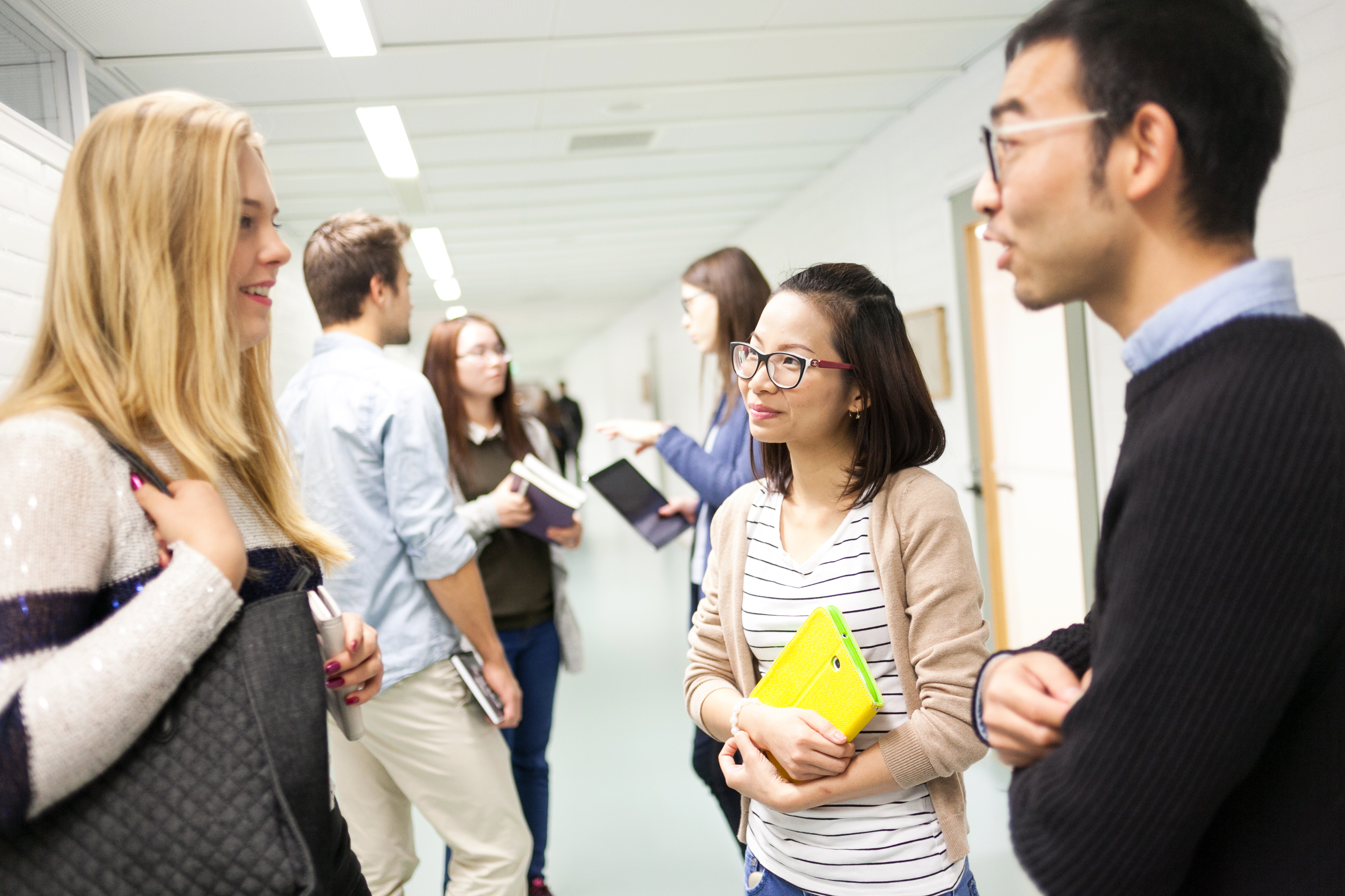 Several students, women and men discussing in the school hallway. One of the women is holding a yellow book.