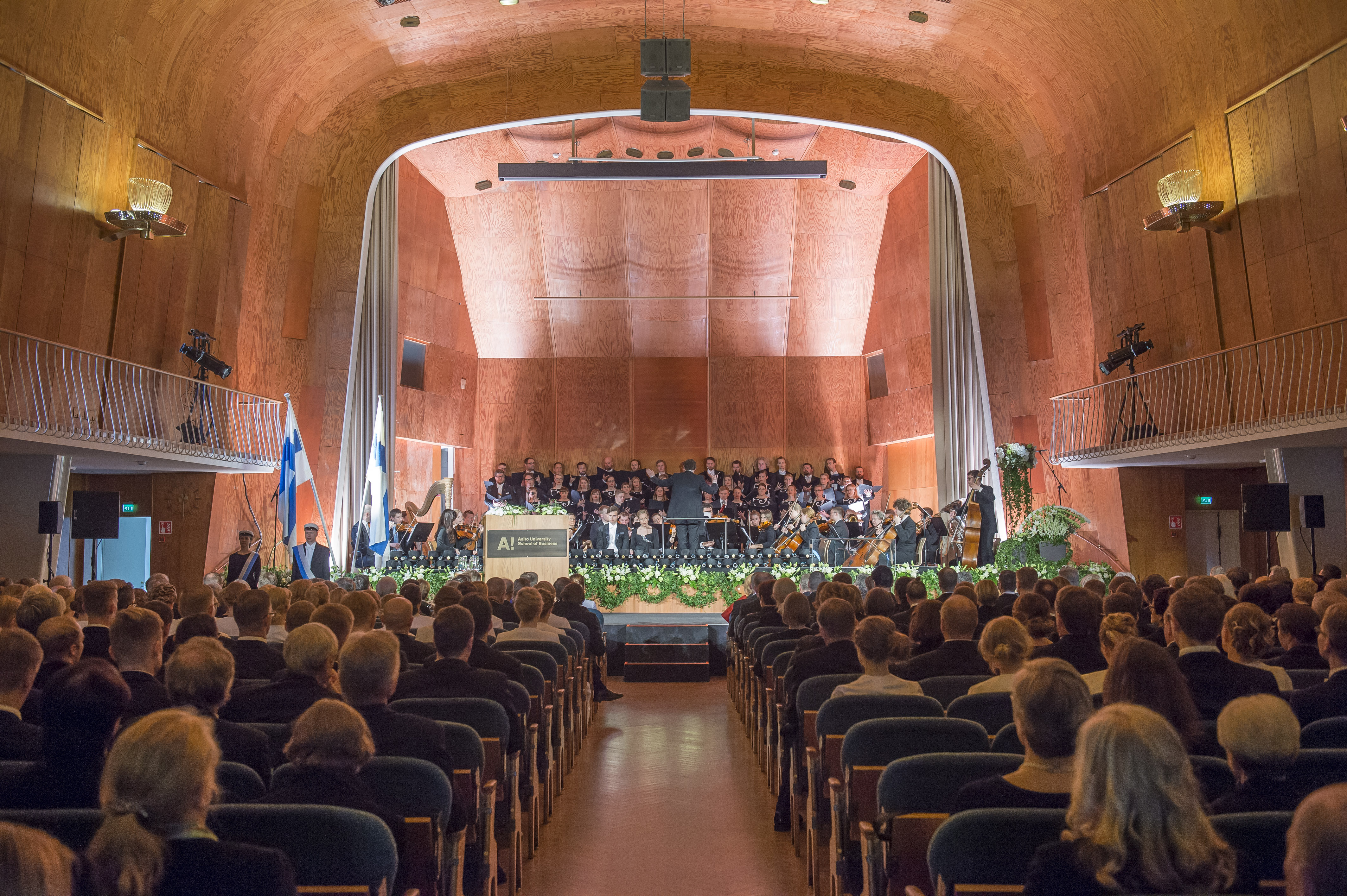 Kuva Kauppakorkeakoulun promootioaktista 2016 Juhlasalissa. Kuvassa yleisöä ja sinfoniaorkesteri kukitetulla lavalla