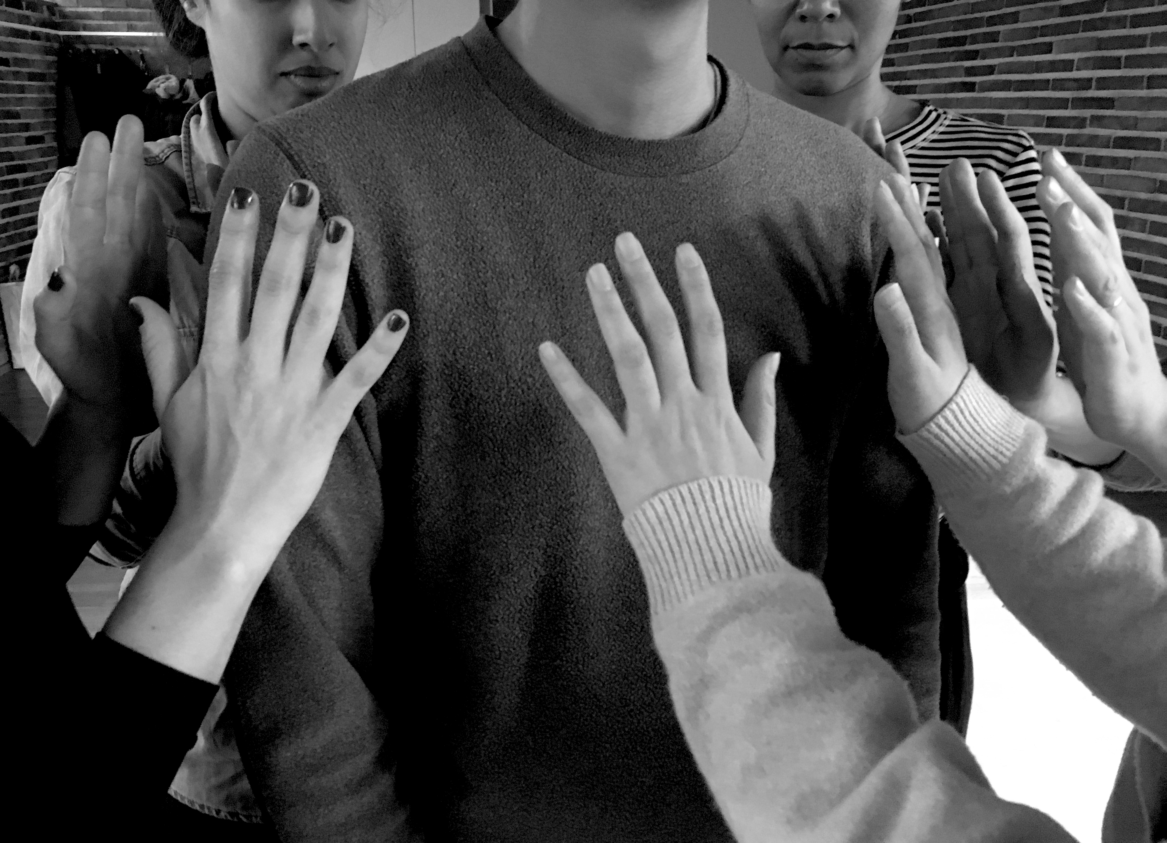 An image from the workshop Situated Knowledges and Embodied Practice. Participants stand in a circle with their hand raised, open, close, to an individual's chest. Greyscale image
