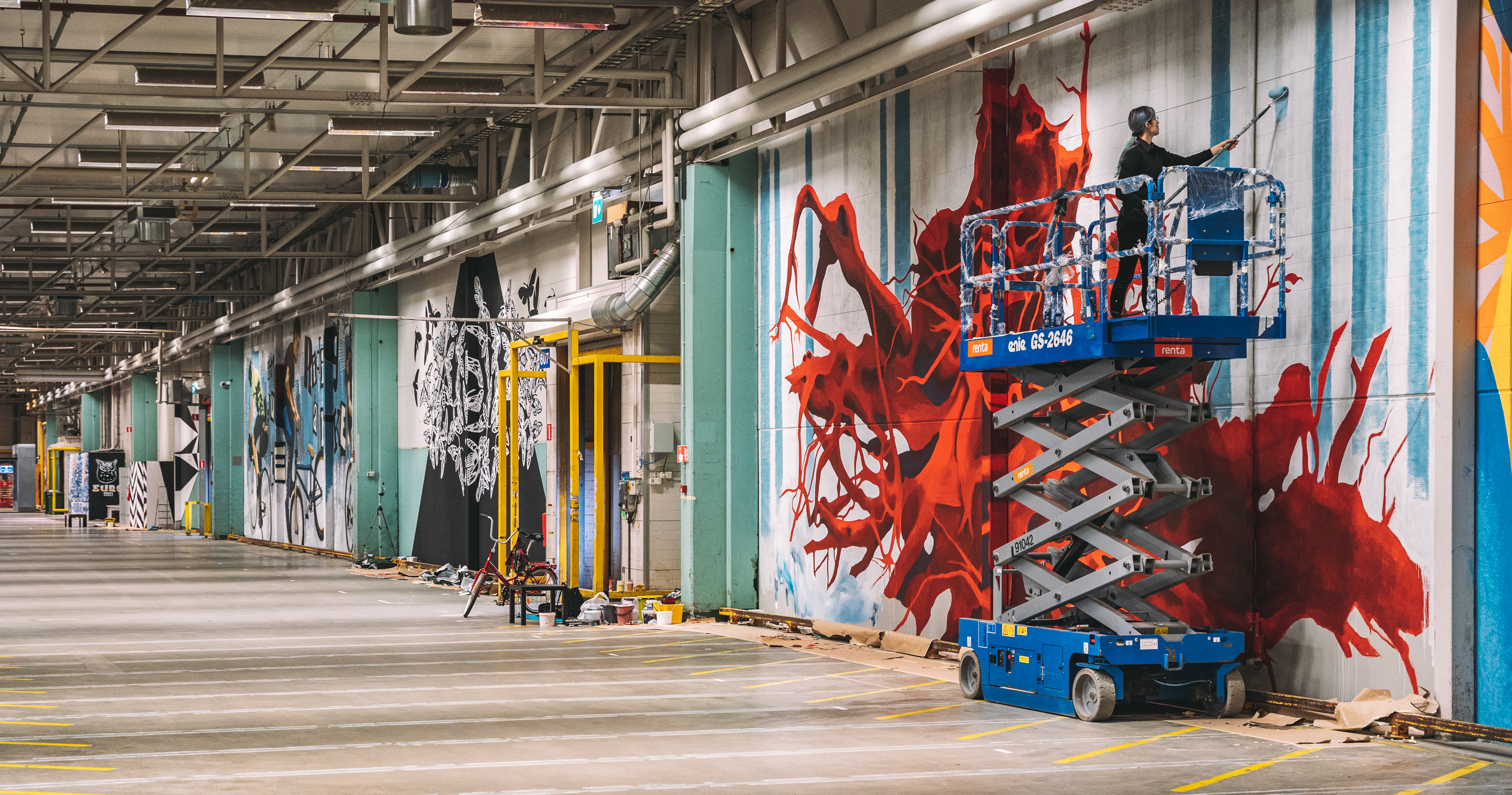 Image of Concreate artists installing their work in Keran Hallit. Artist Saara Salmi stands on a scissor-lift to reach her painting