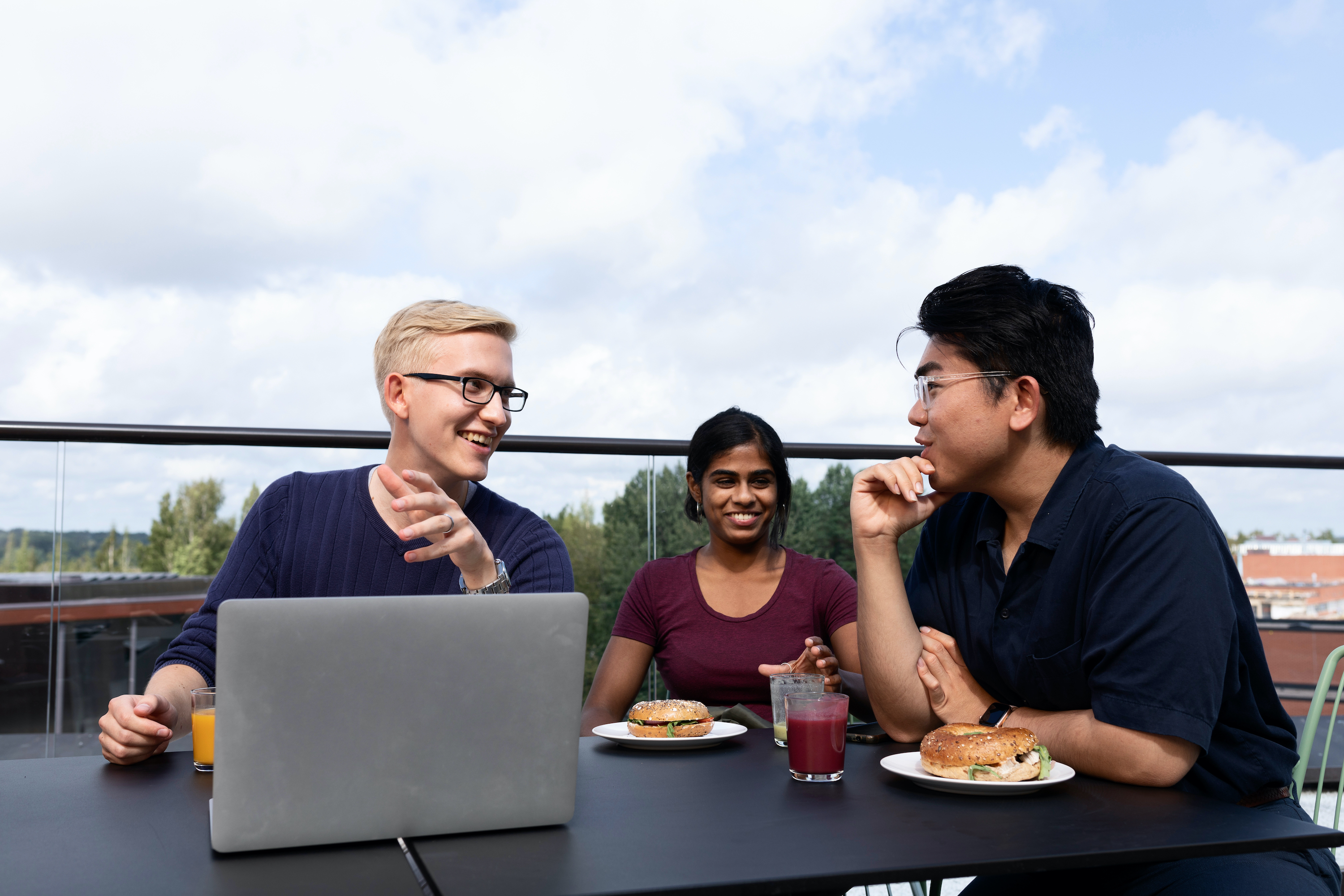 Three students eating a snack and discussing over a computer on a rooftop terrace. 