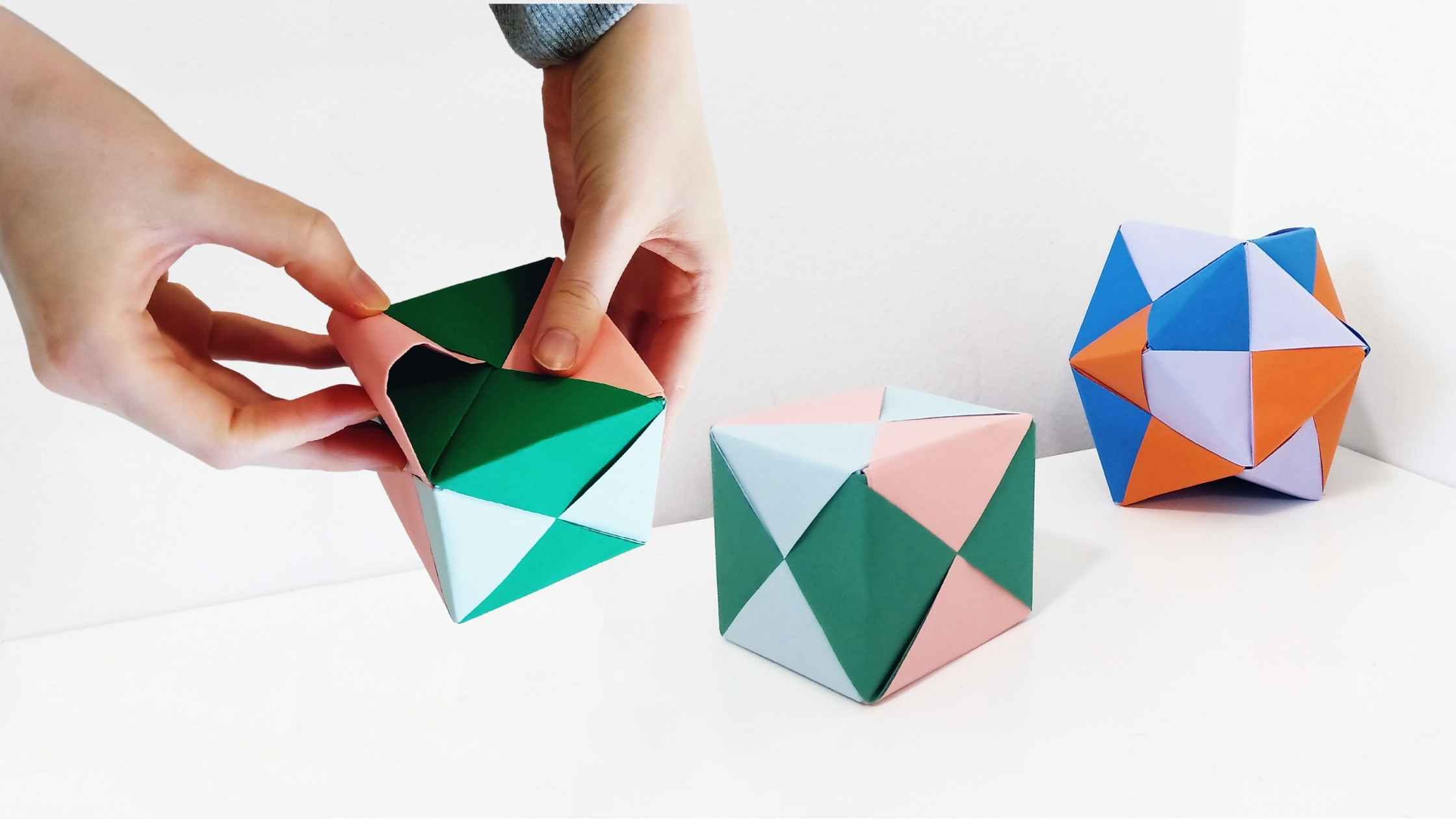 Kädet taittelevat värikästä origamikuuutiota, taustalla origamikuutio sekä tähtimäistetty oktaedri