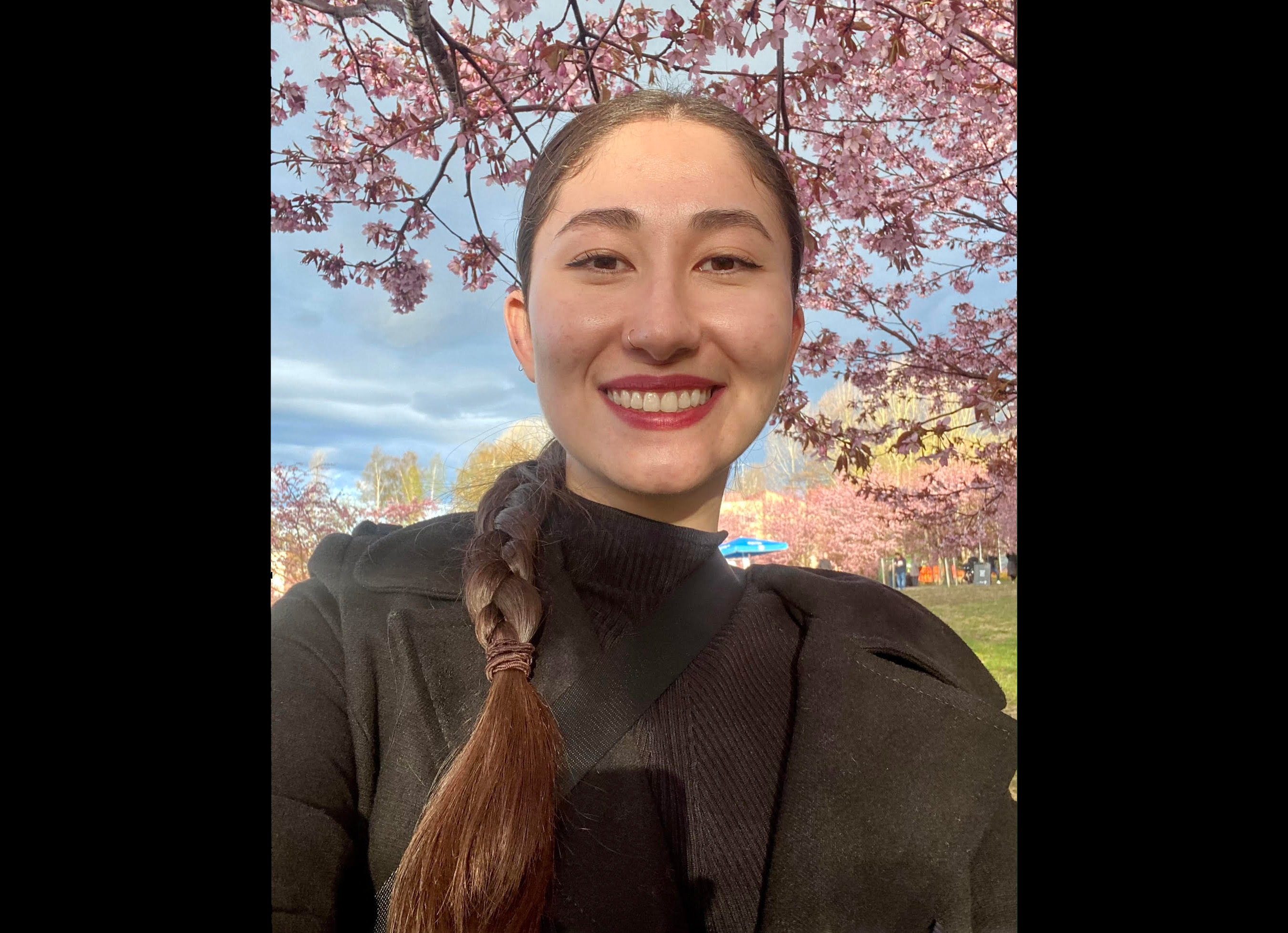 Person with braided hair, wearing a black jacket under cherry blossom trees.