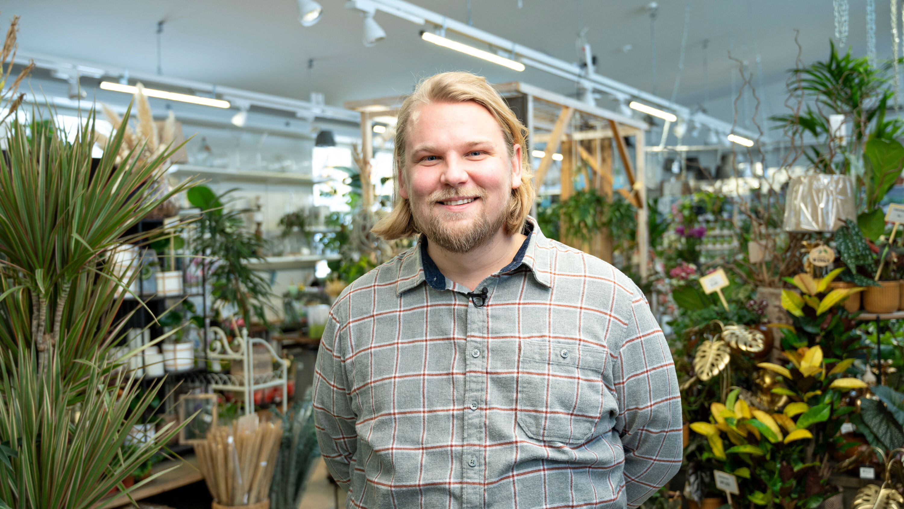 A person wearing a grey checked shirt is standing in a store filled with various plants and gardening supplies.
