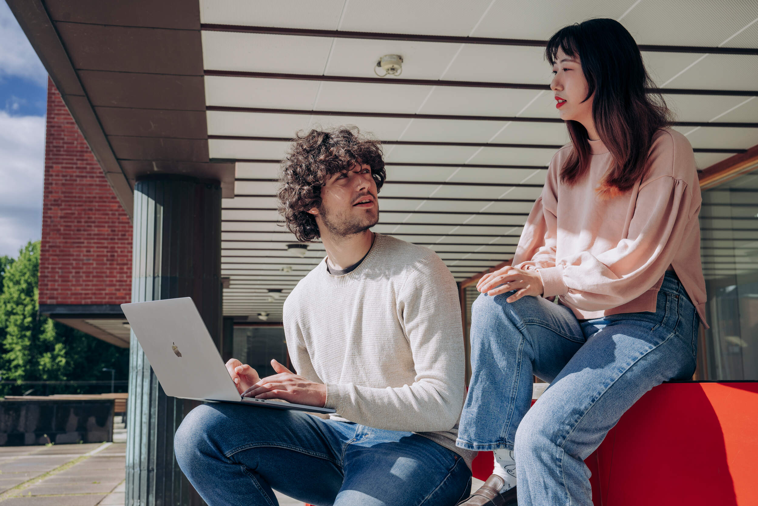Two students sitting and discussing outside while one student is holding a laptop in his knee while listening to the other student.