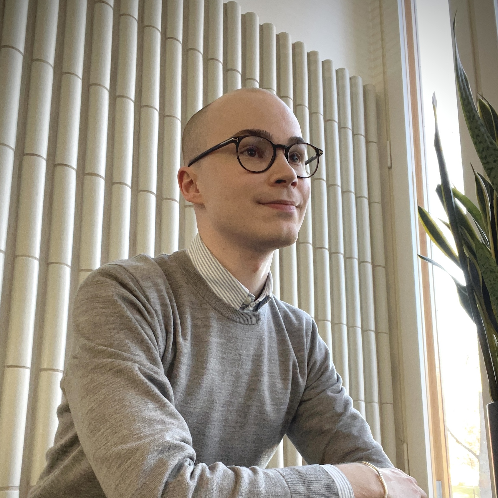 A PhD researcher with dark glasses sits near a window on the Aalto University campus.