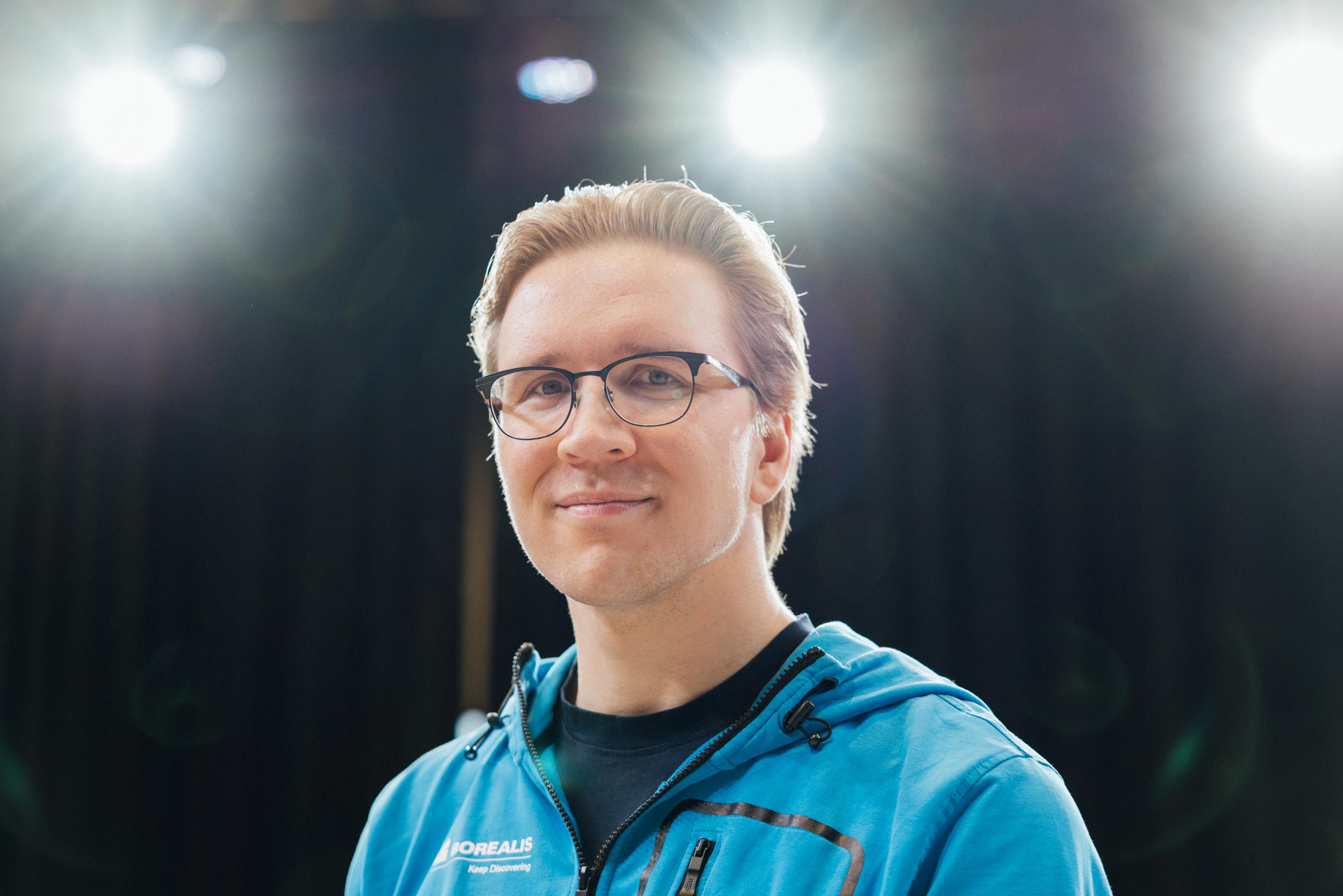 Henkilö sinisessä takissa kirkkaiden valojen alla, musta tausta.