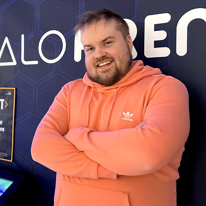 Oranssiin huppariin pukeutunut henkilö seisoo käsivarret ristissä seinän edessä, jossa on geometrisia kuvioita ja tekstiä.