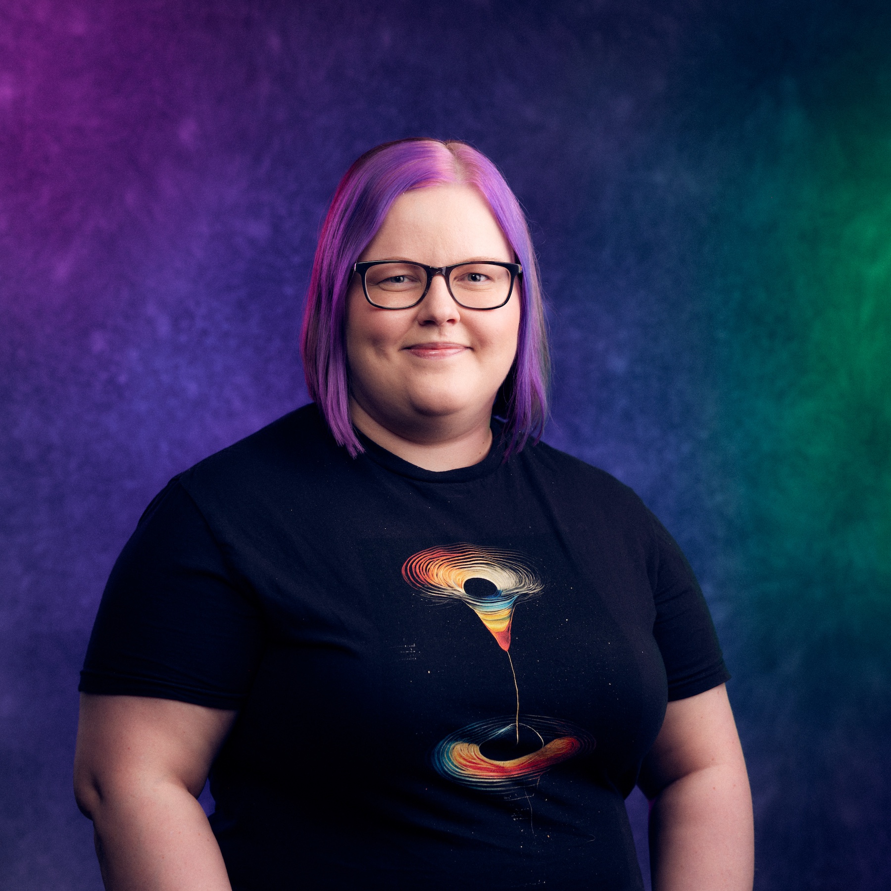 A person with shoulder-length hair wearing a black shirt featuring a colourful abstract design. The background is a gradient of purple, blue, and green.