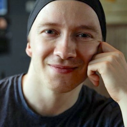 Henkilö, jolla on tumma paita ja hattu, istuu sisätiloissa ja nojaa kämmenselkäänsä.