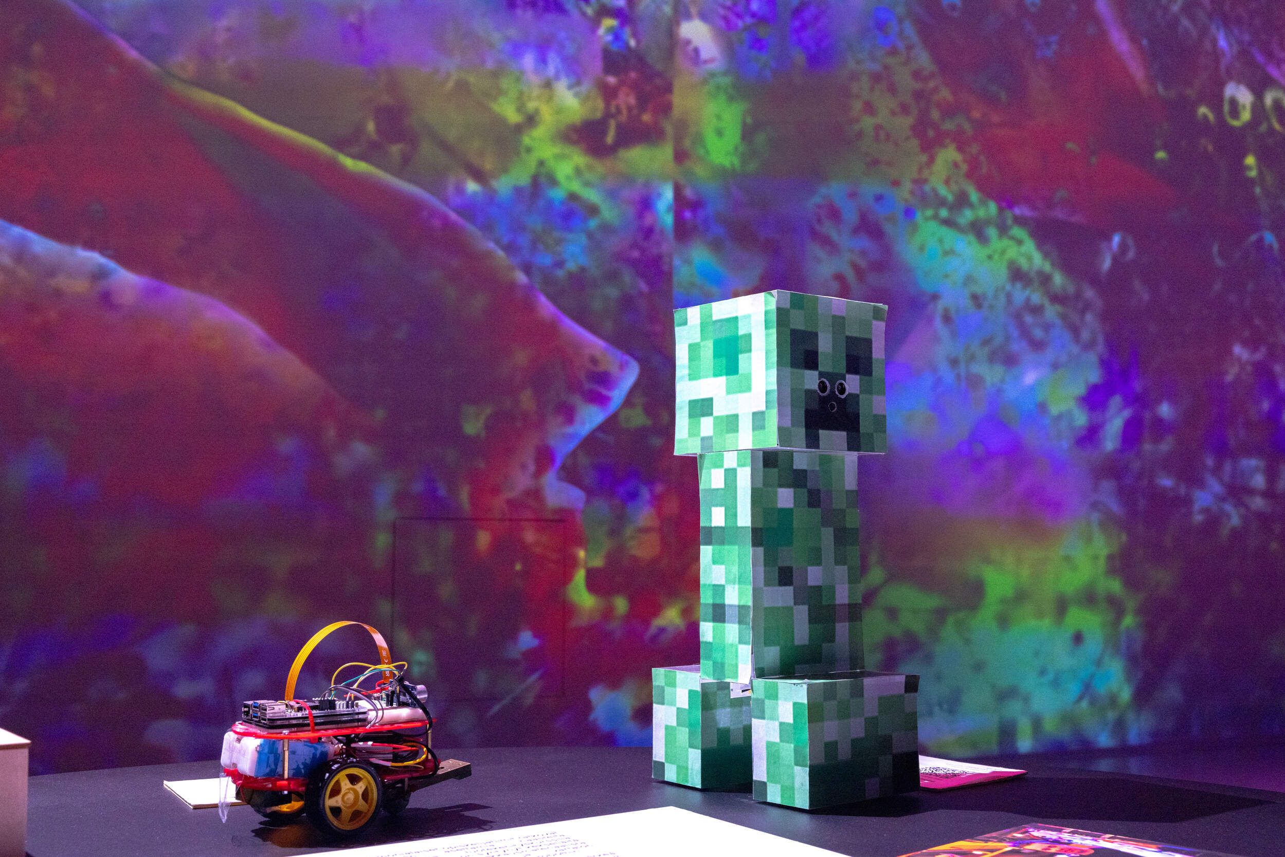 Minecraft Creeper -hahmo seisoo pienen pyörillä varustetun robotin vieressä värikkään taustan edessä.