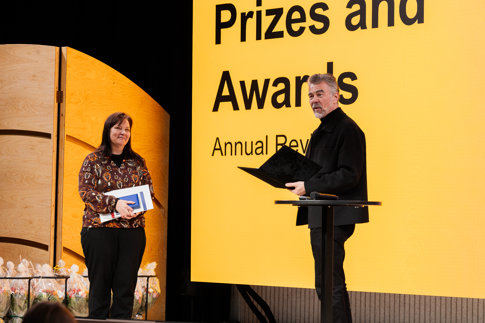 Two people on stage at an awards ceremony. One holds a folder; the other holds a book with a background marked 'Prizes and Awards'.