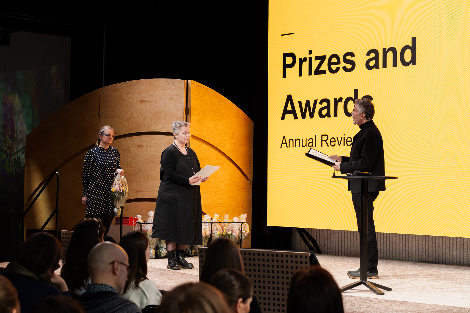 A prize ceremony with two women receiving awards from a man in a black outfit. Yellow background with text.