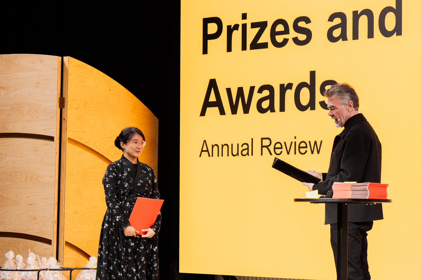 Two people at an awards ceremony, one holding a red folder and the other black. Yellow wall with the text 'Prizes and Awards'.