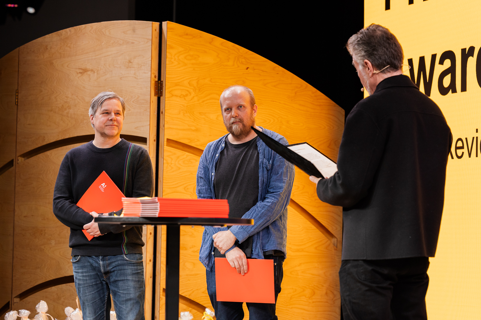 Three people onstage holding red folders, one is speaking. Wooden backdrop and sign reading 'award' visible.