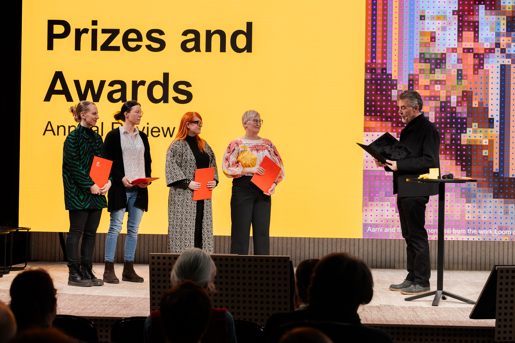 Four people on stage holding red certificates, with a man presenting an award. Background shows 'Prizes and Awards'.