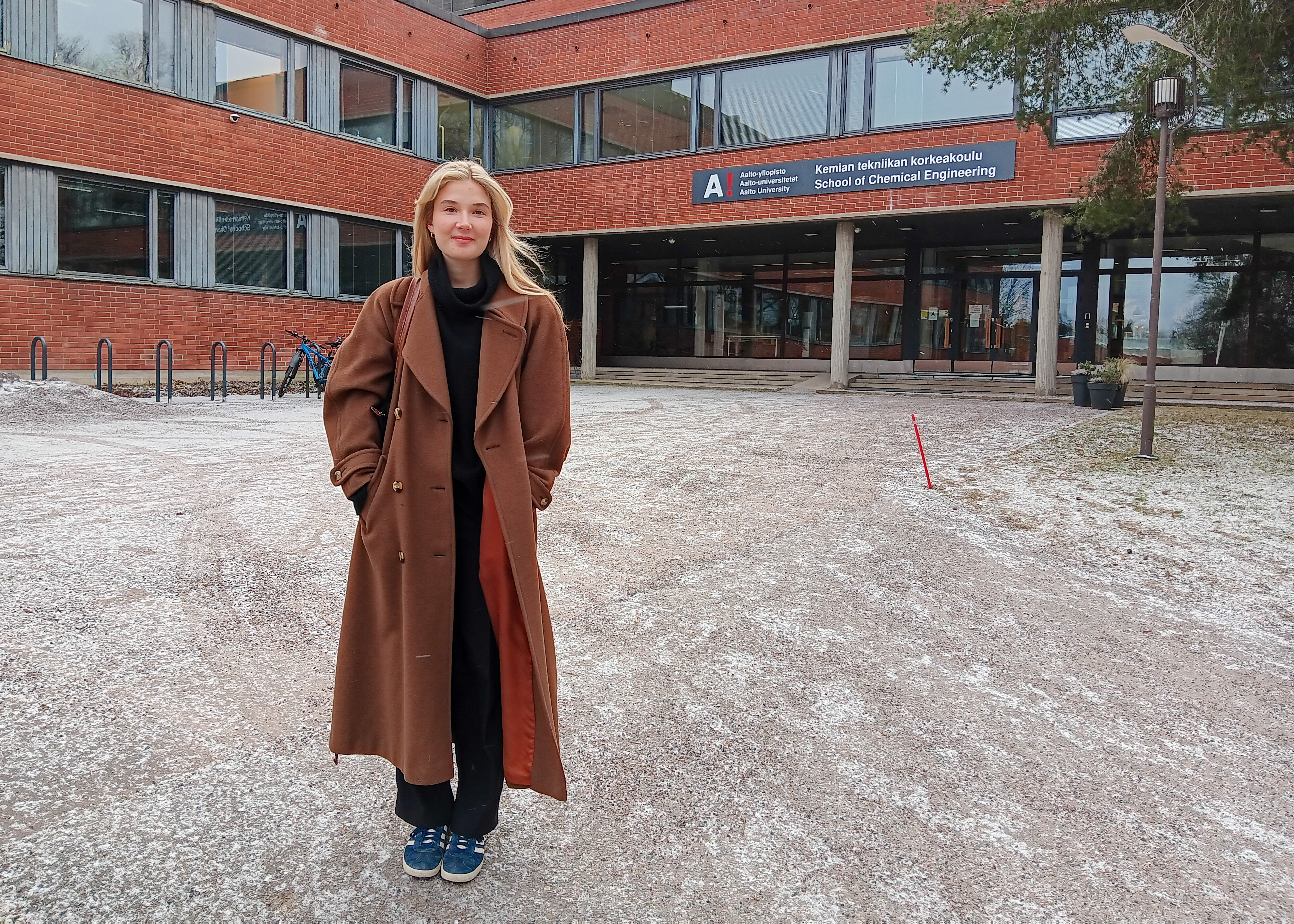 Henkilö pitkässä ruskeassa takissa seisoo rakennuksen edessä, jossa on 'Kemian tekniikan korkeakoulu' -kyltti.