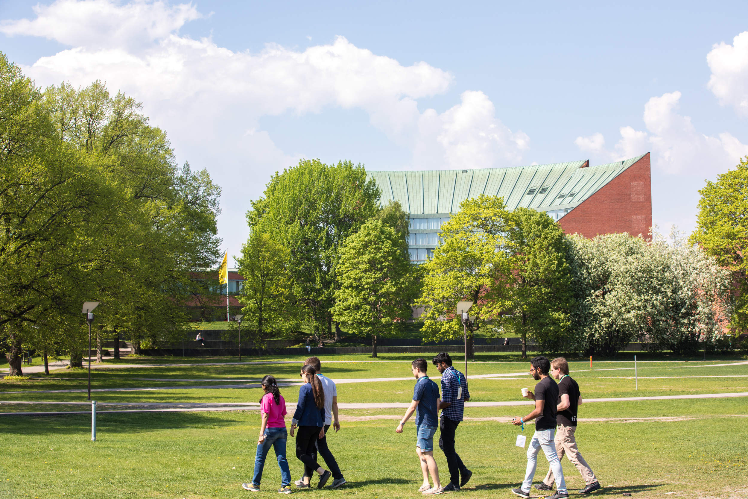Group of people walking across a green park with trees. Aalto's buildings in the background.