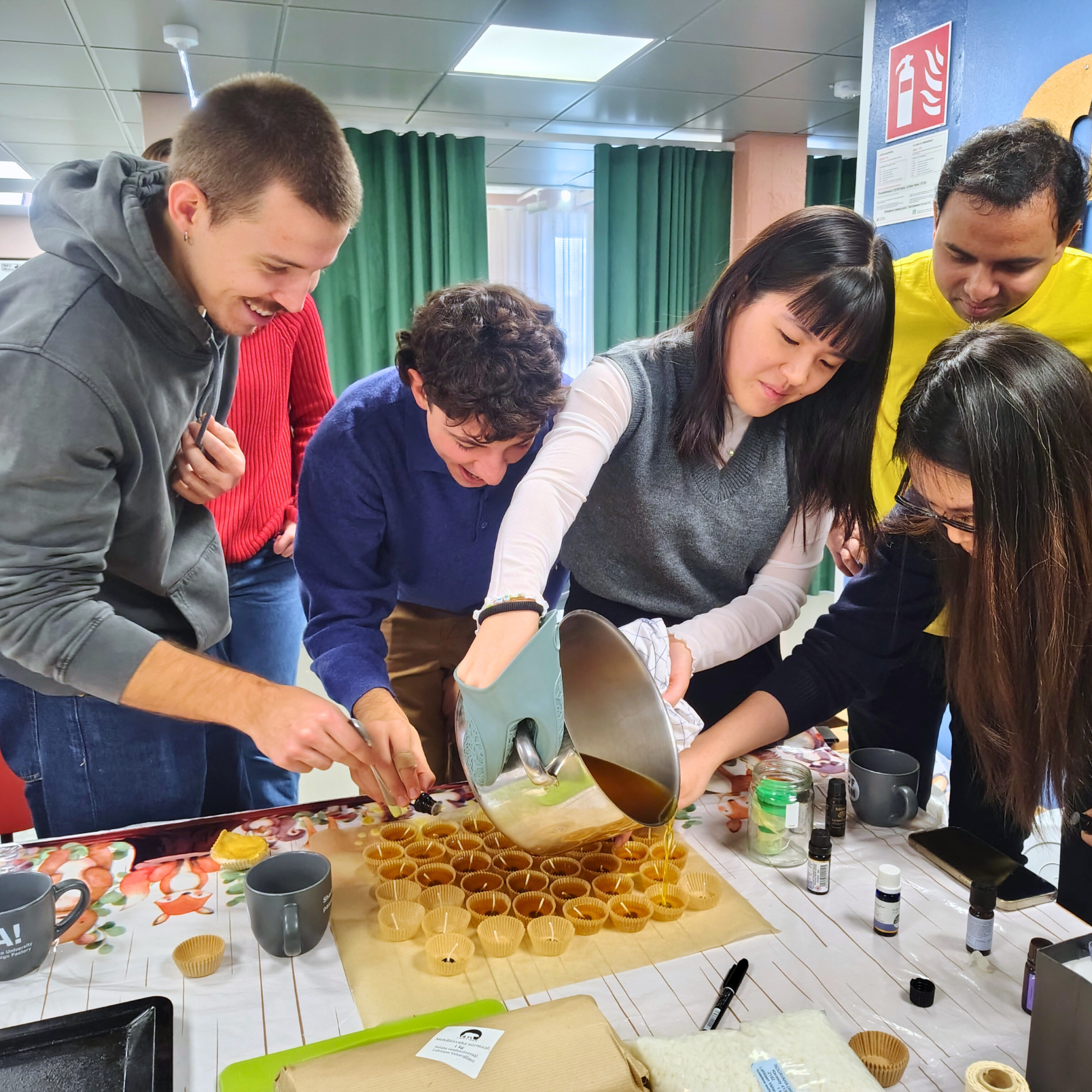 A group of people making candles from bees wax. One person is pouring wax into moulds on a table covered with various items.