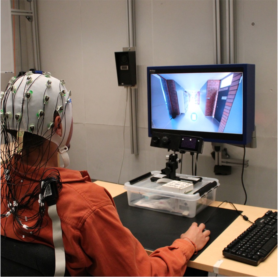 A person wearing an EEG cap sits at a computer, viewing a display with a virtual corridor.