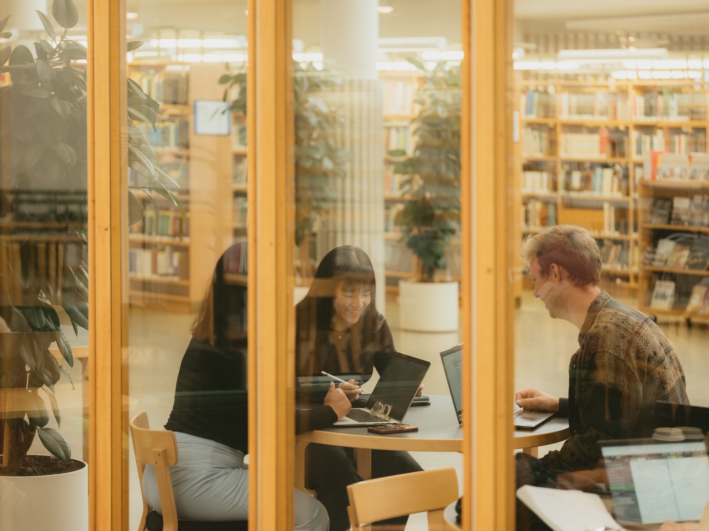 Three people working on laptops at a round table in a library, seen through glass walls.
