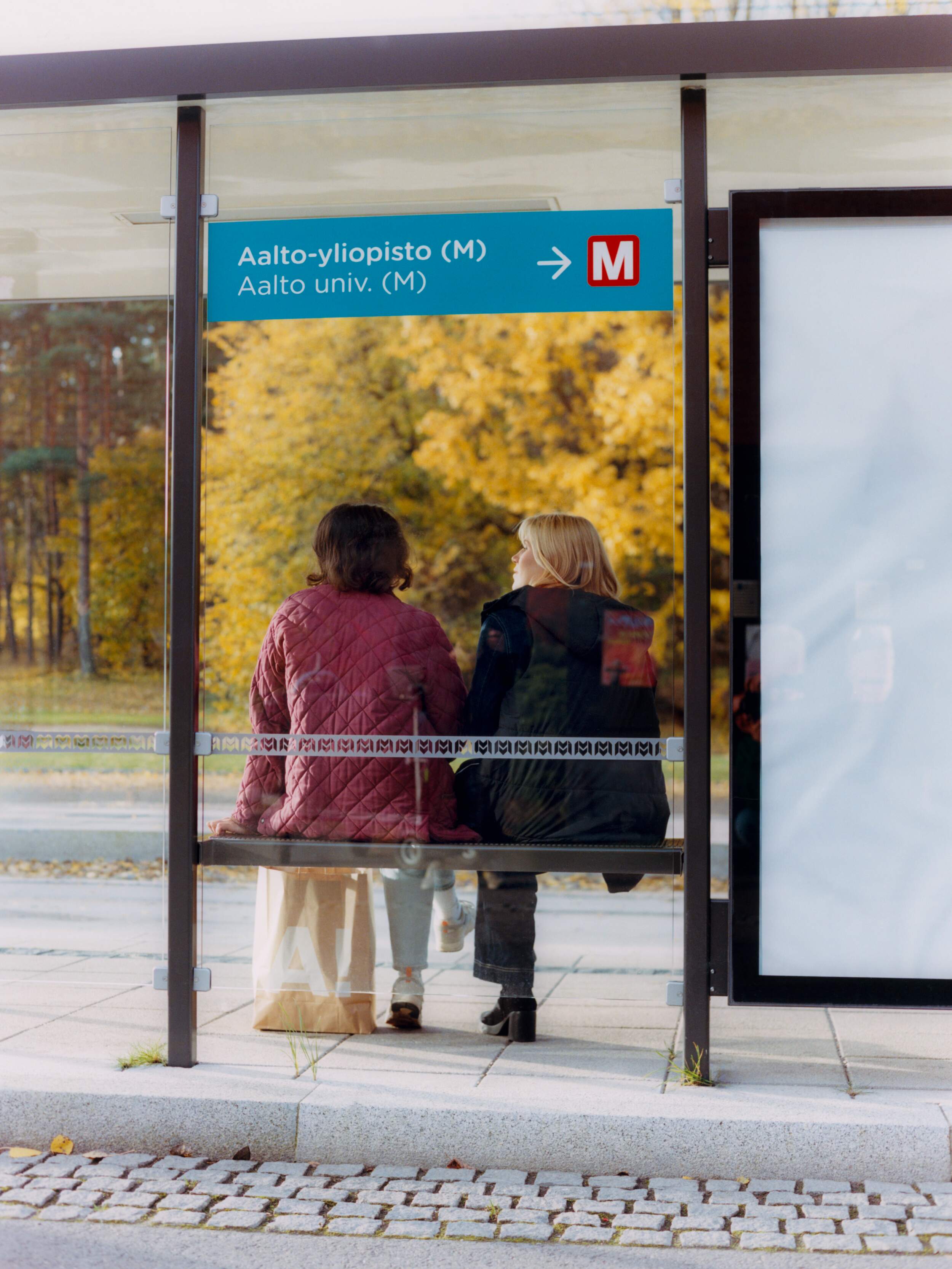 Two people sitting at a bus stop with a sign for Aalto University. Autumn trees are visible in the background.
