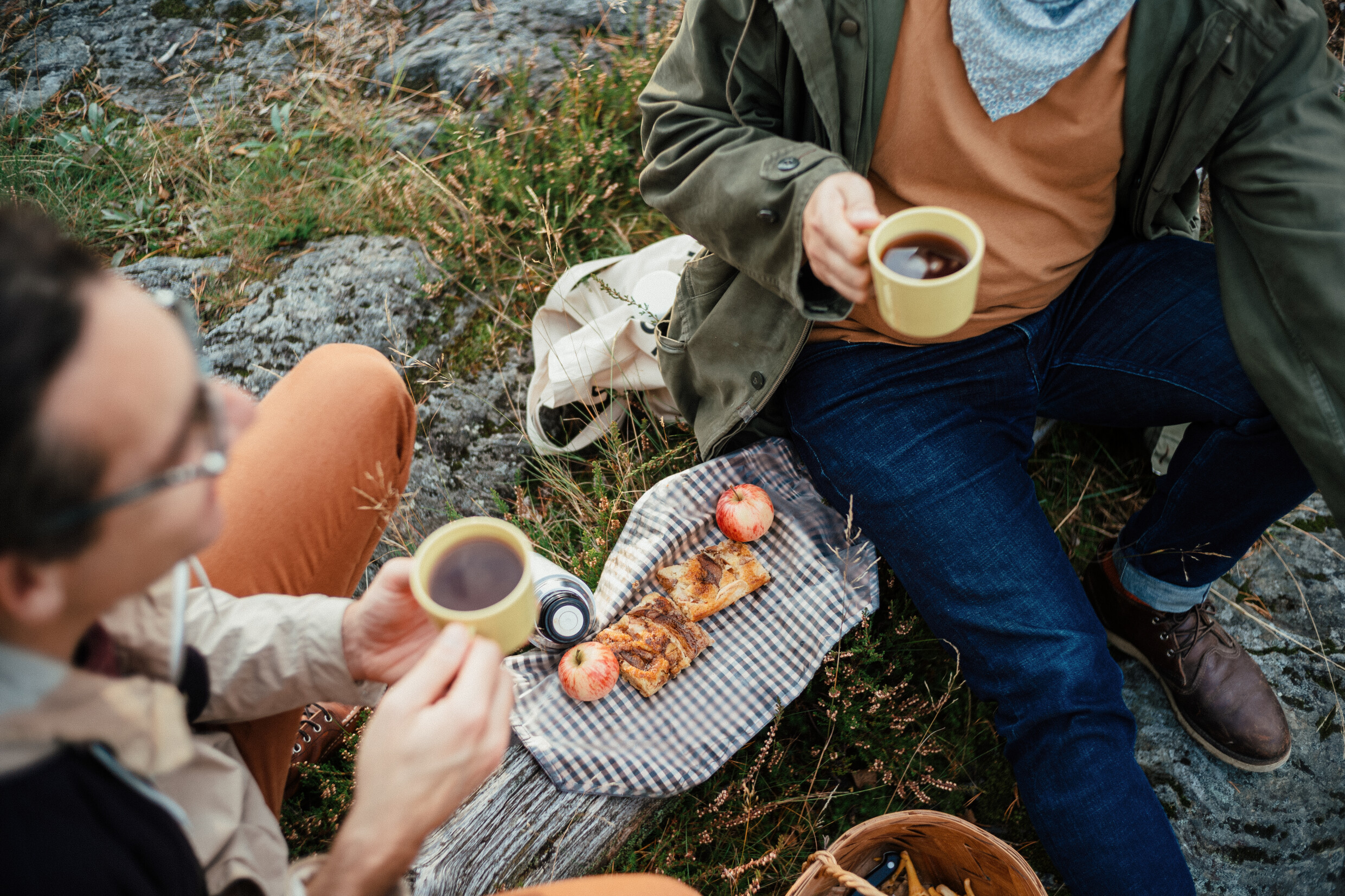 Two people picnicking outdoors, holding coffee mugs. On the cloth are pastries, an apple, and a camera.