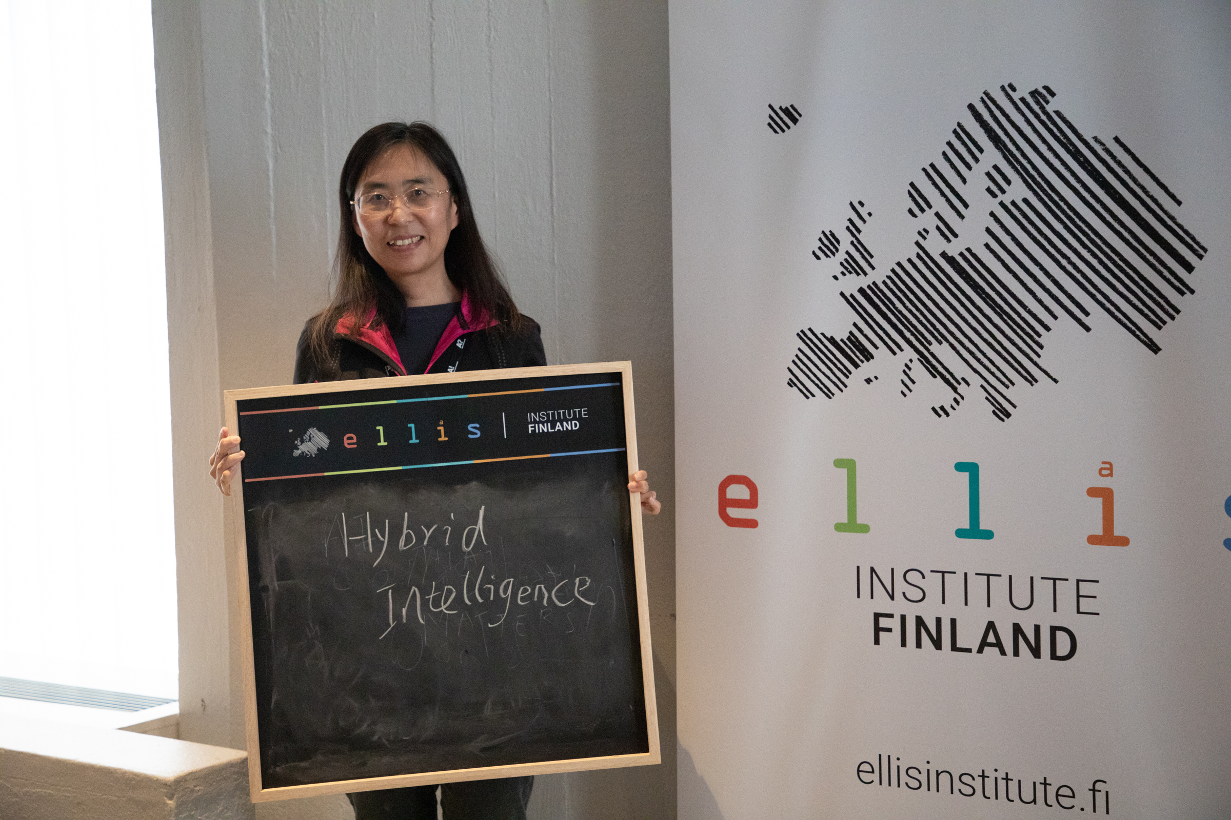 A person holds a chalkboard with 'Hybrid Intelligence' written on it in front of an ELLIS Institute Finland banner.