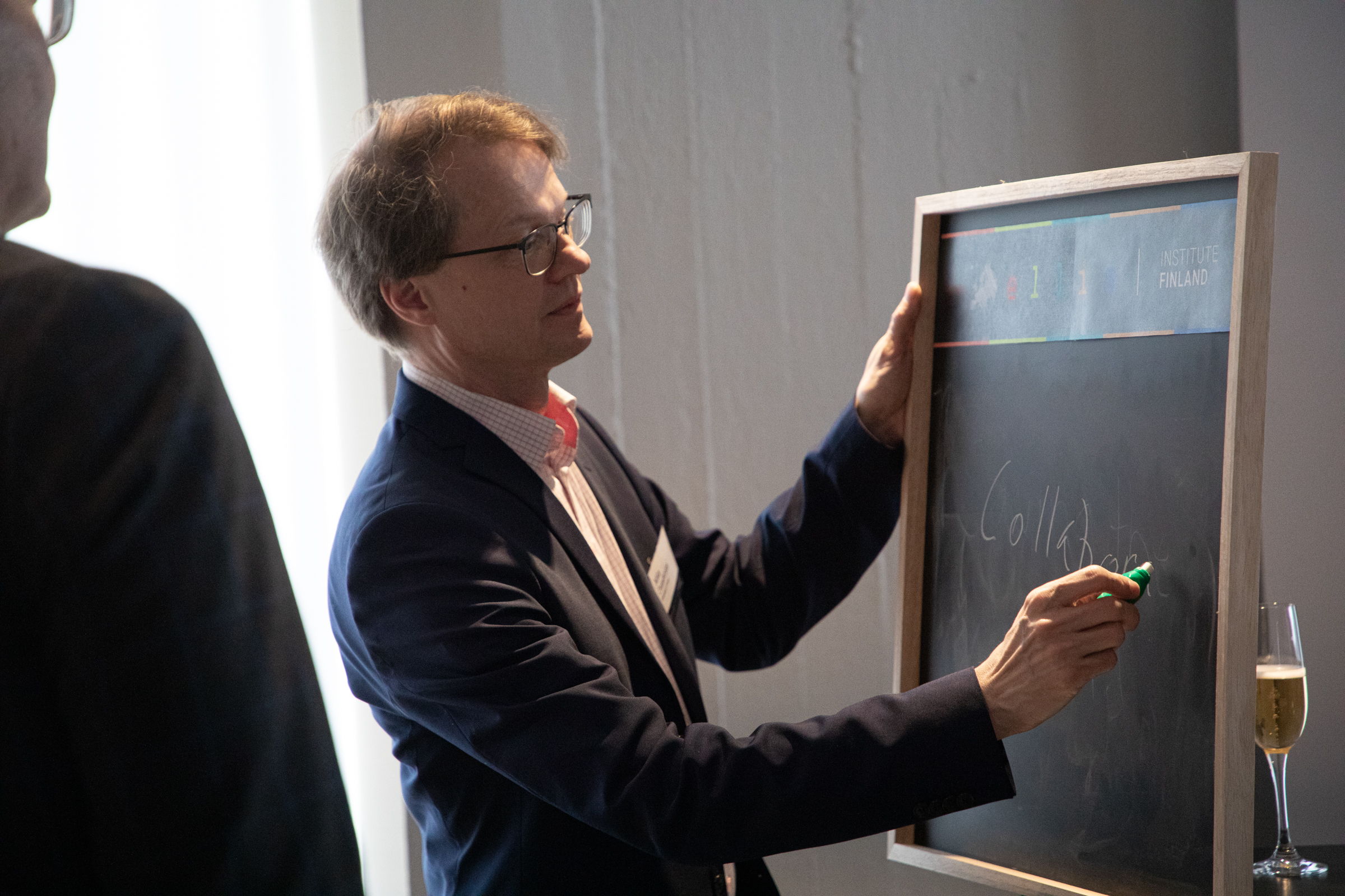 A man holding a chalkboard and writing with a green chalk. A glass of champagne is next to him.