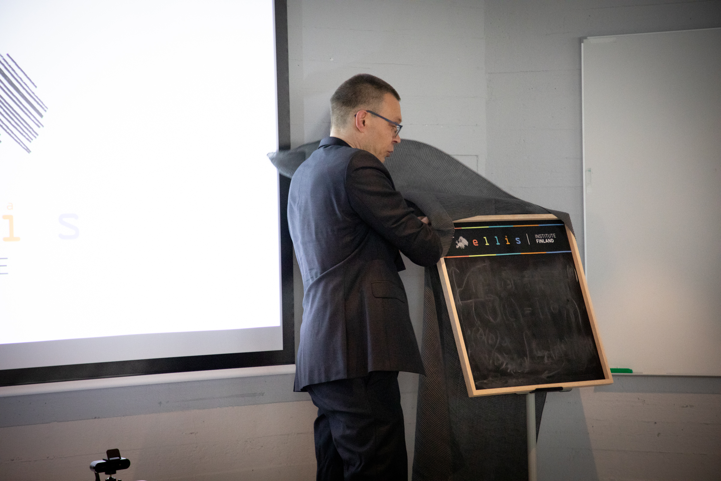 Man in a suit unveiling a blackboard next to a large whiteboard and projector screen.