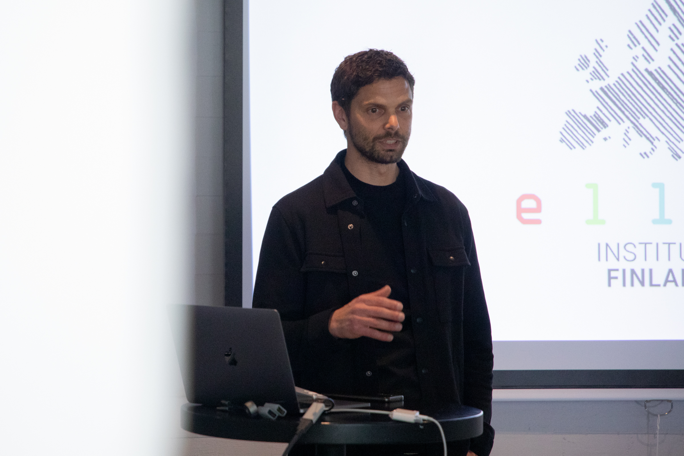 A person wearing a black outfit gives a presentation with a laptop on a table. A large screen shows text on the right.