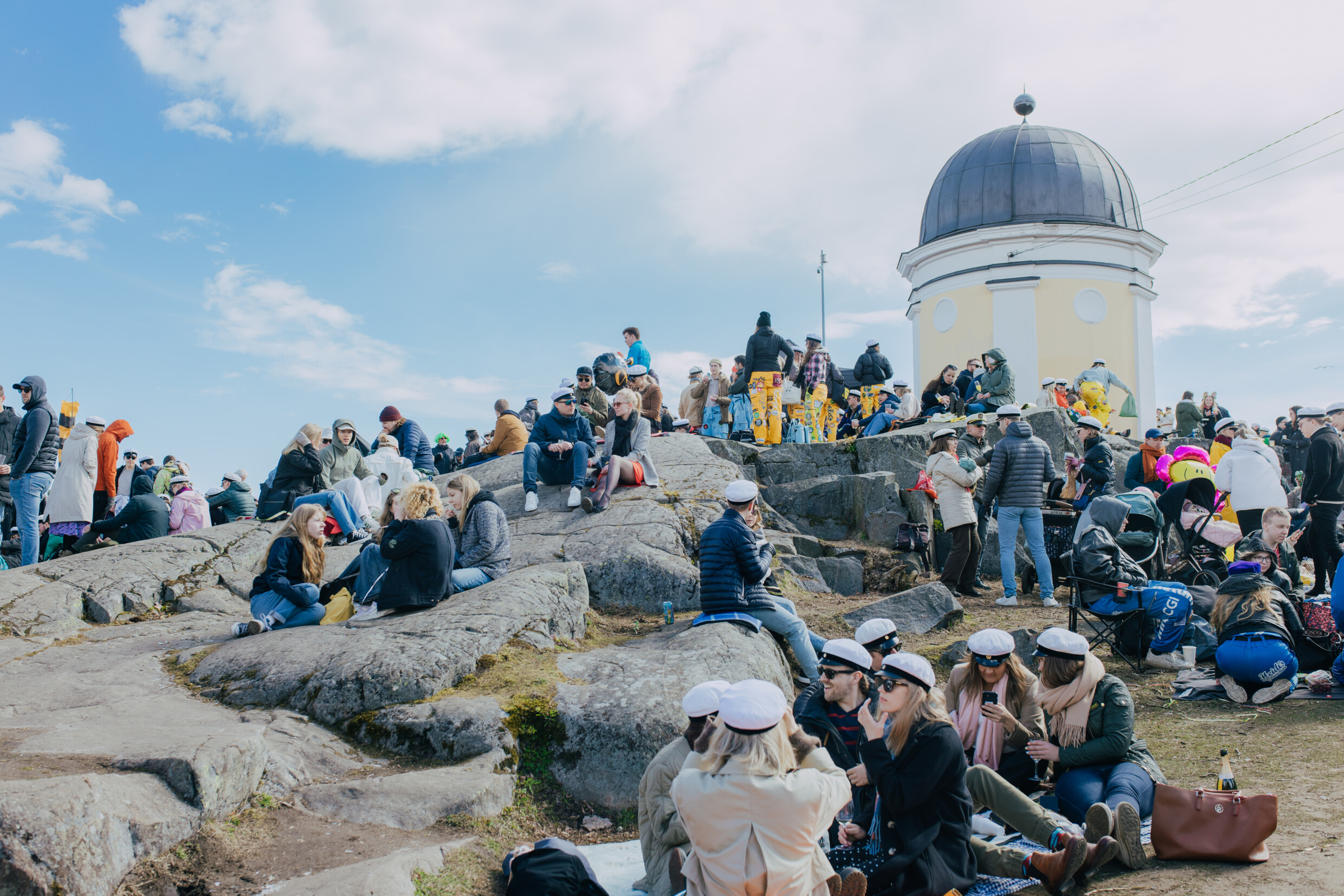 A large group of students celebrating outdoors on rocky terrain near a yellow observatory tower, many wearing white caps.
