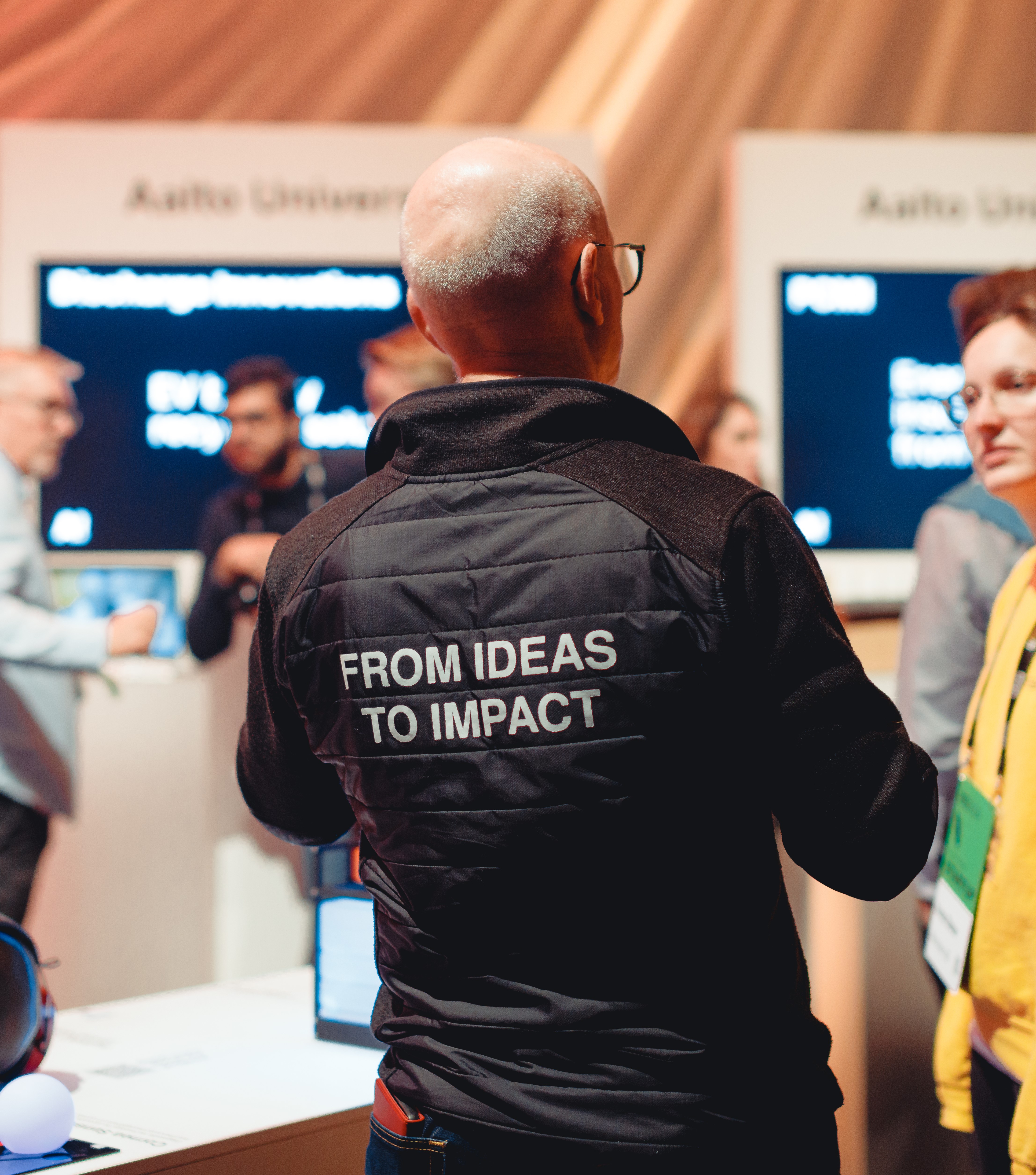 Henkilö mustassa takissa, jossa valkoinen teksti 'FROM IDEAS TO IMPACT'. Yliopistotapahtuma, taustalla sumeita ihmisiä.