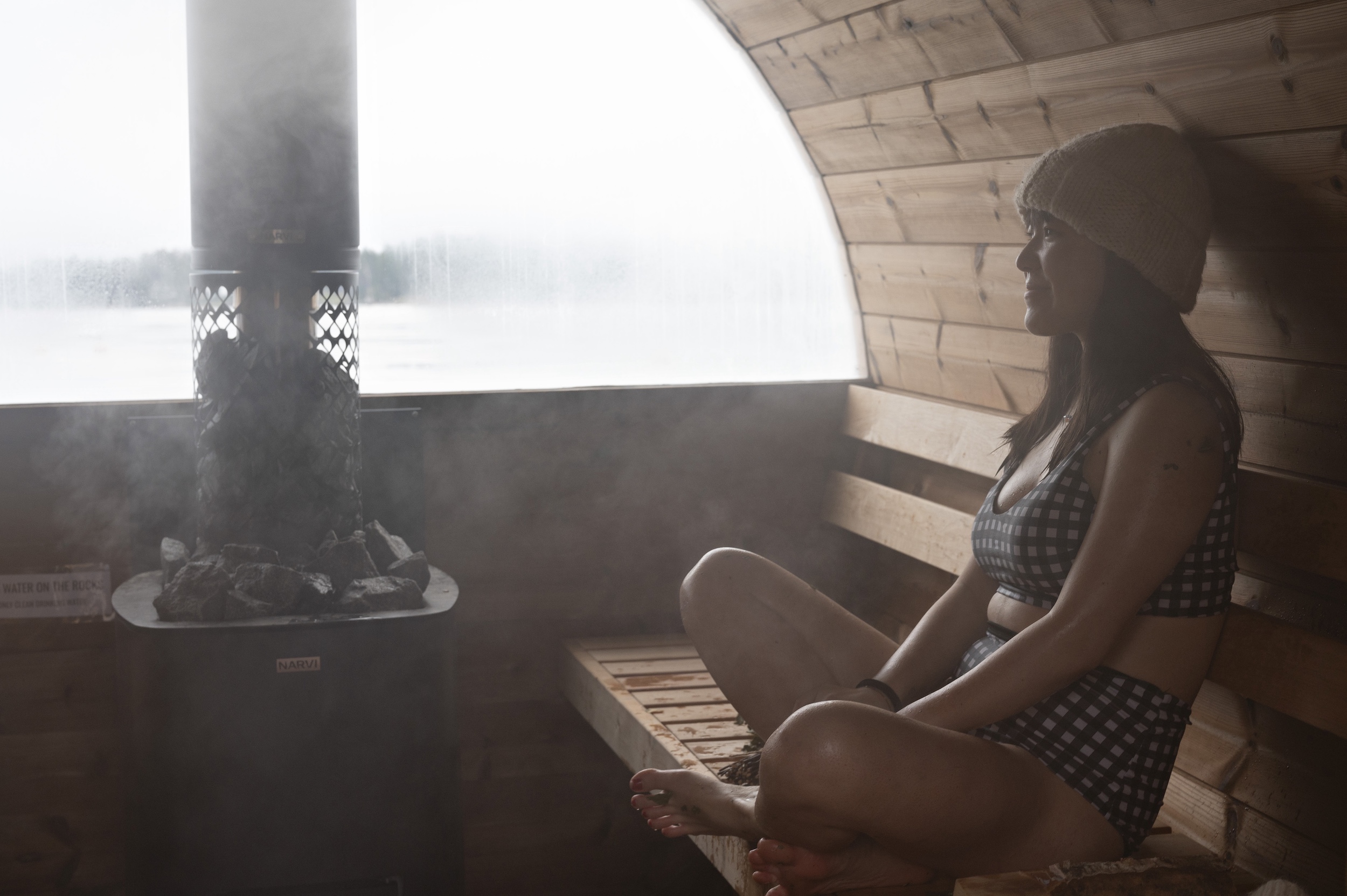 A woman sits in a wooden sauna wearing a checked two-piece swimsuit and a beanie, with a stove and a misty window in the background.