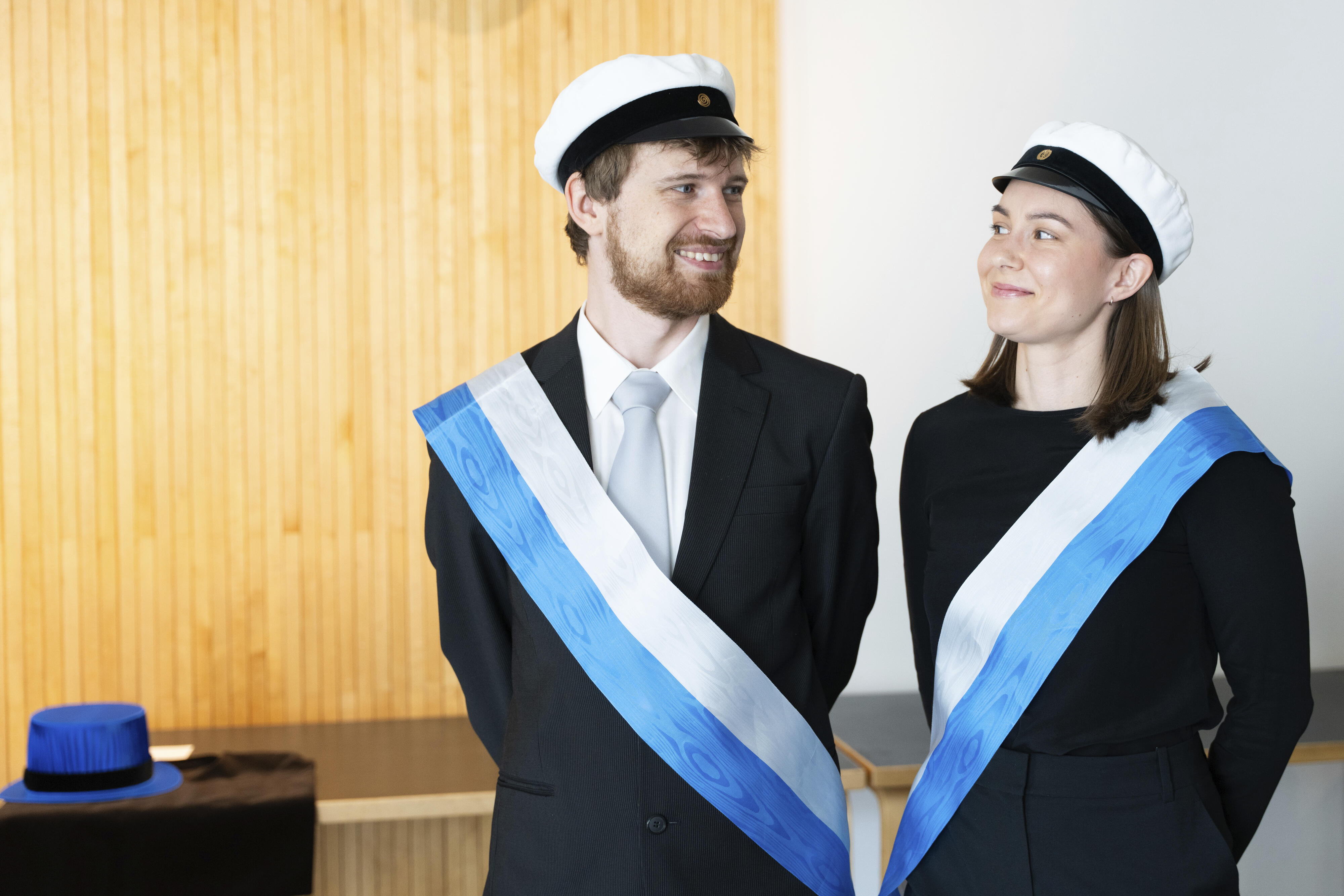 Kaksi henkilöä mustissa juhla-asuissa ja valkolakeissa, valkoiset ja siniset nauhat, seisovat sisällä.
