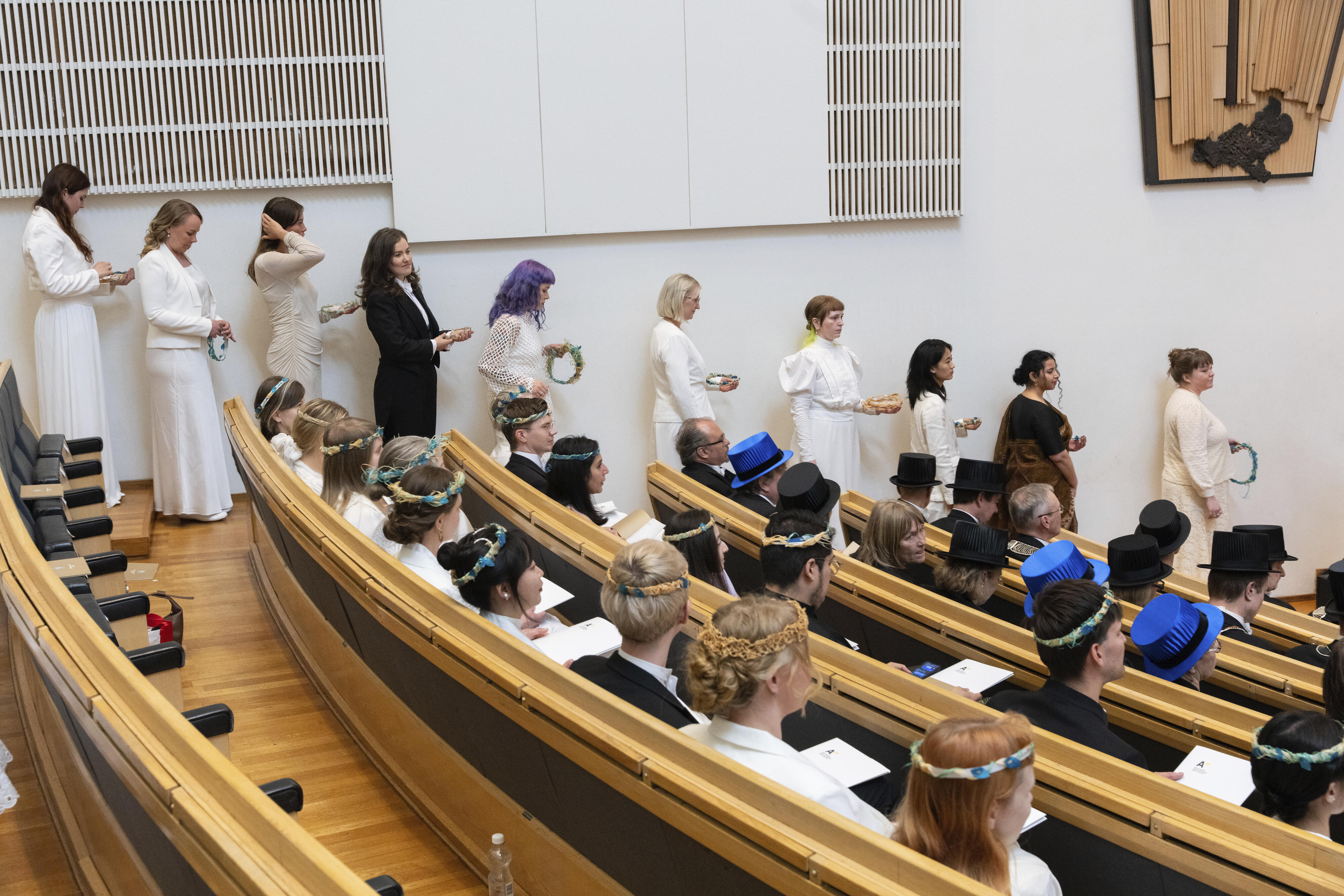 Ihmiset valkoisissa akateemisissa kaavuissa ja puvuissa jonottavat luentosalissa. Osa käyttää laakeriseppeleitä ja akateemisia lakkeja.