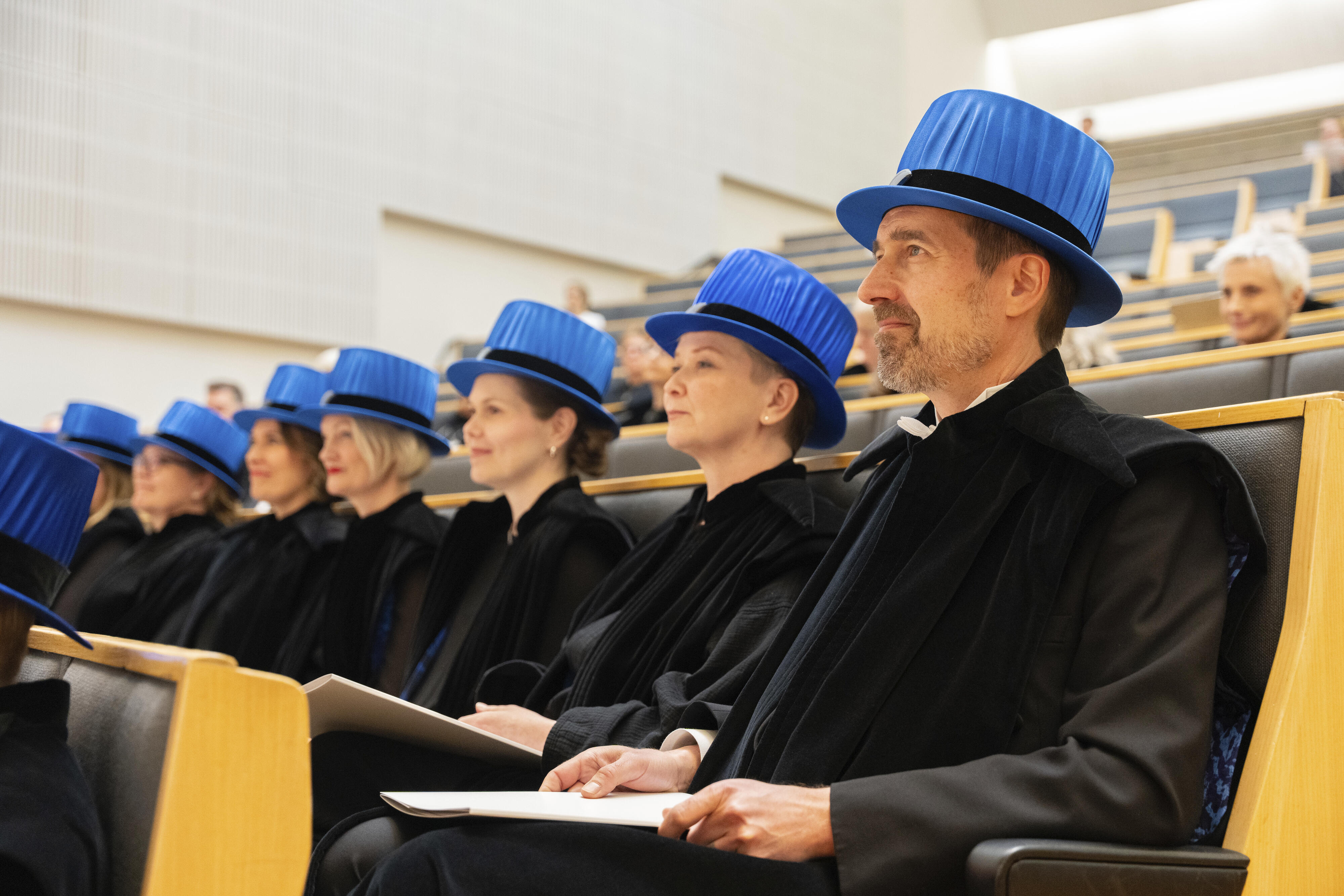Valmistuneet tummissa kaavuissa ja sinisissä hatuissa istuvat luentosalissa pitäen diplomeja.