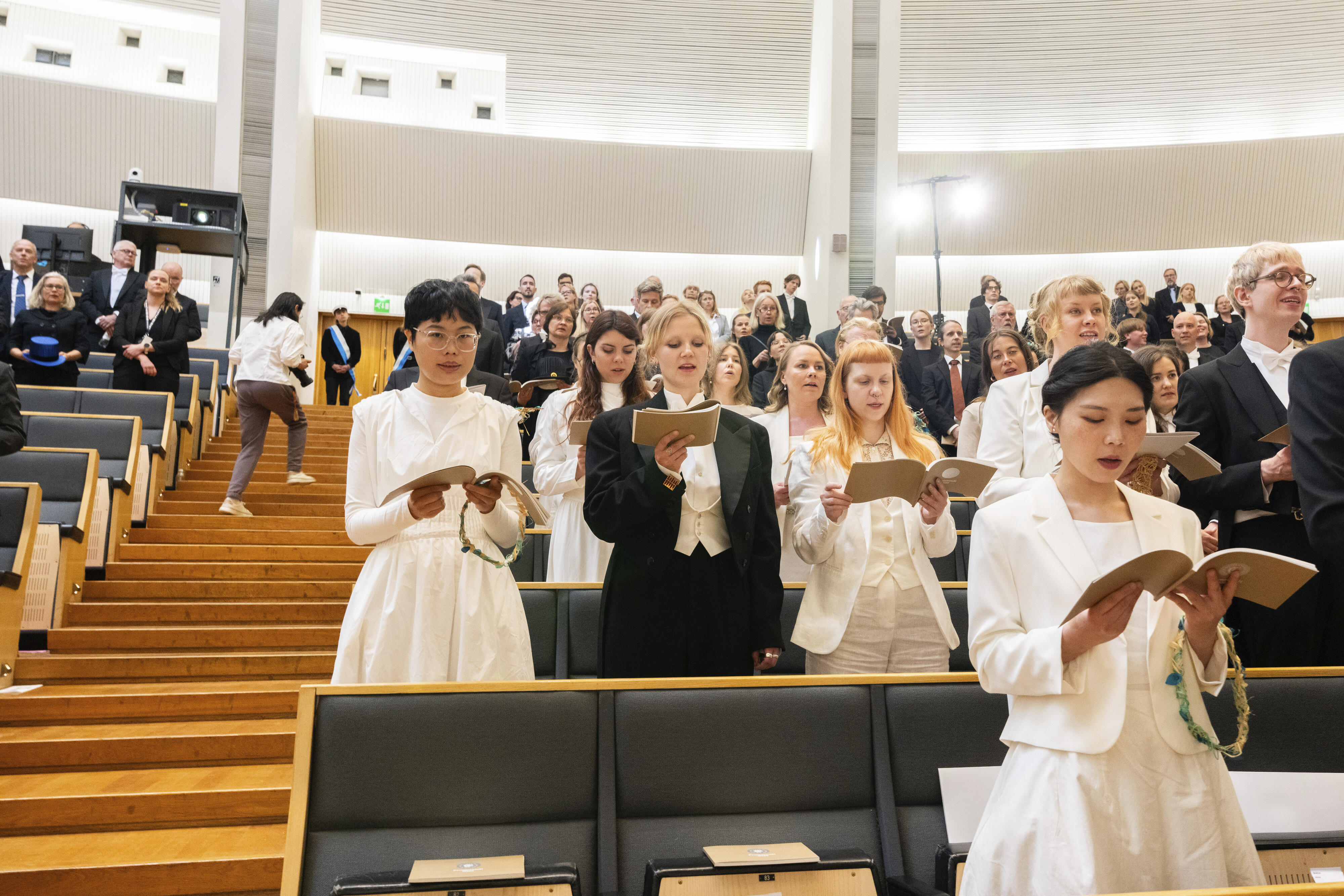 Ihmiset pukeutuneet juhlavasti laulavat auditoriossa laulukirjoja kädessä, taustalla seisoo muita ihmisiä.