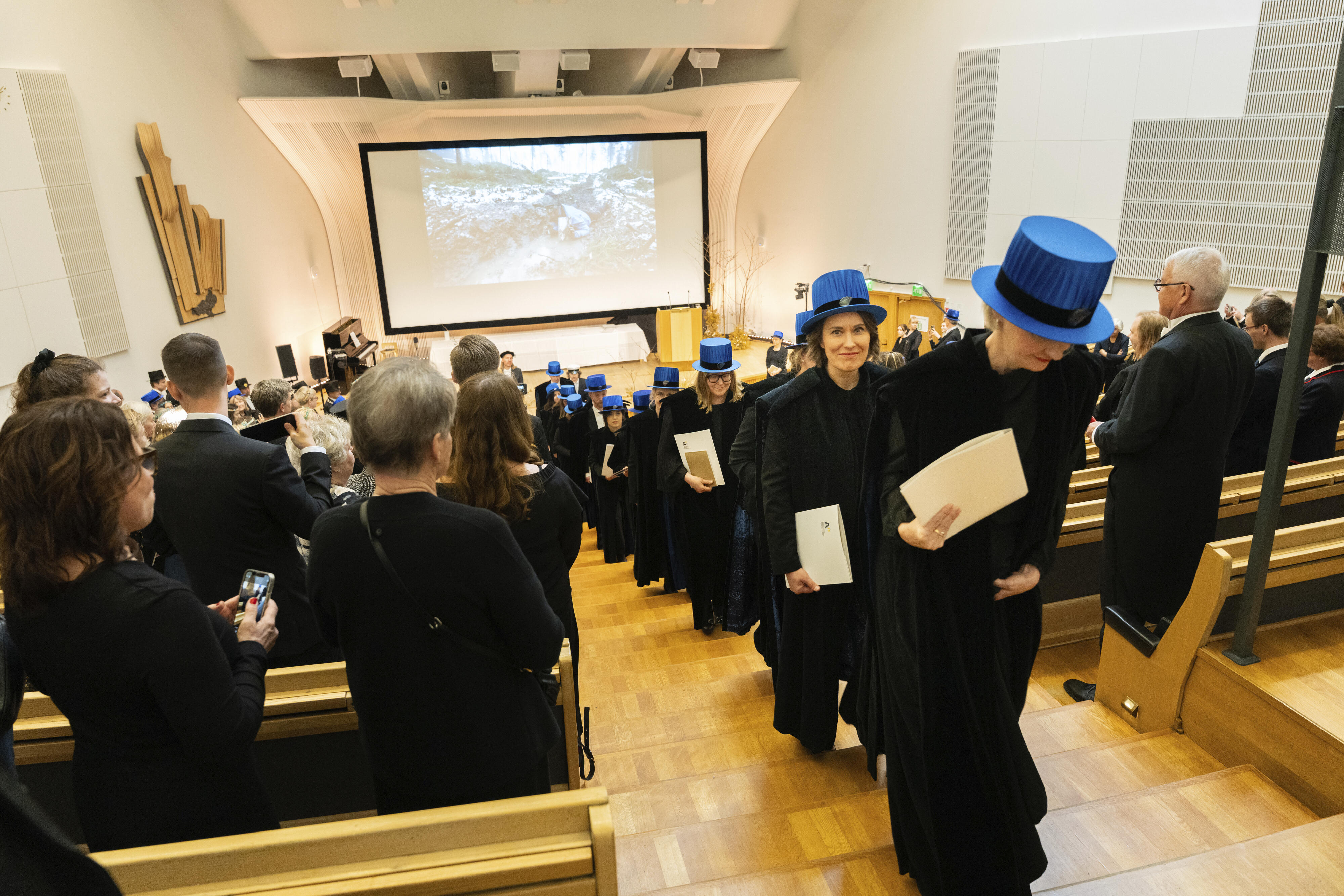 Graduation ceremony in an auditorium, graduates dressed in black gowns and blue hats holding diplomas.