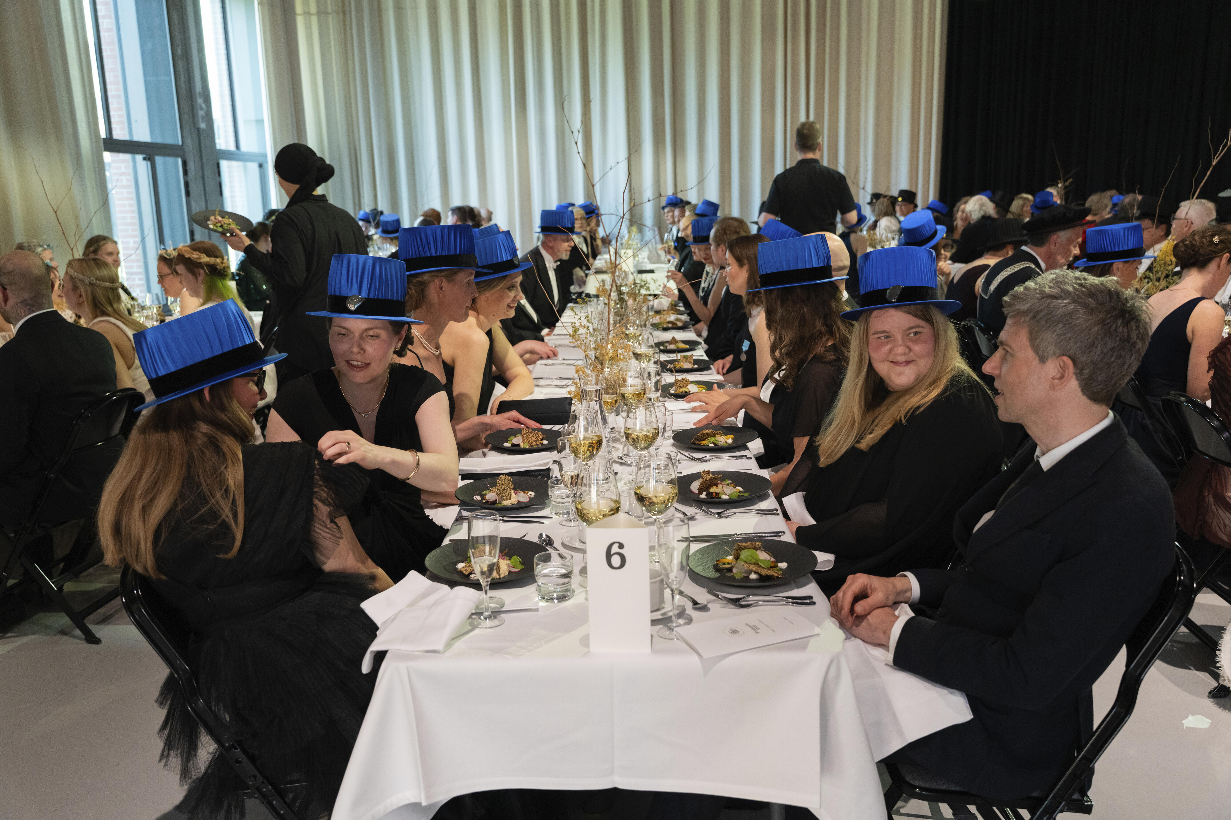 Ihmiset puvuissa ja sinisissä akateemisissa hatuissa istuvat pitkän juhlapöydän ääressä, numero 6 kortti näkyvillä.