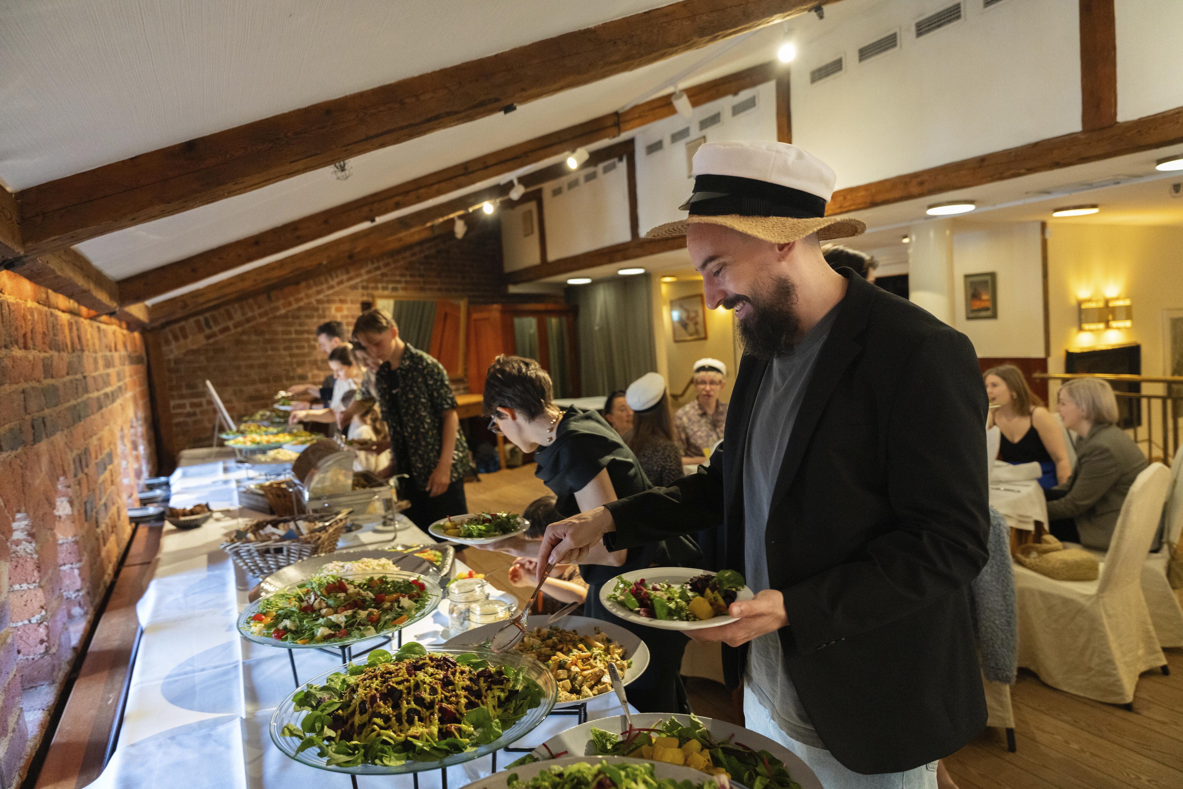 People in casual attire enjoy an indoor buffet. The room has exposed beams and brick walls.