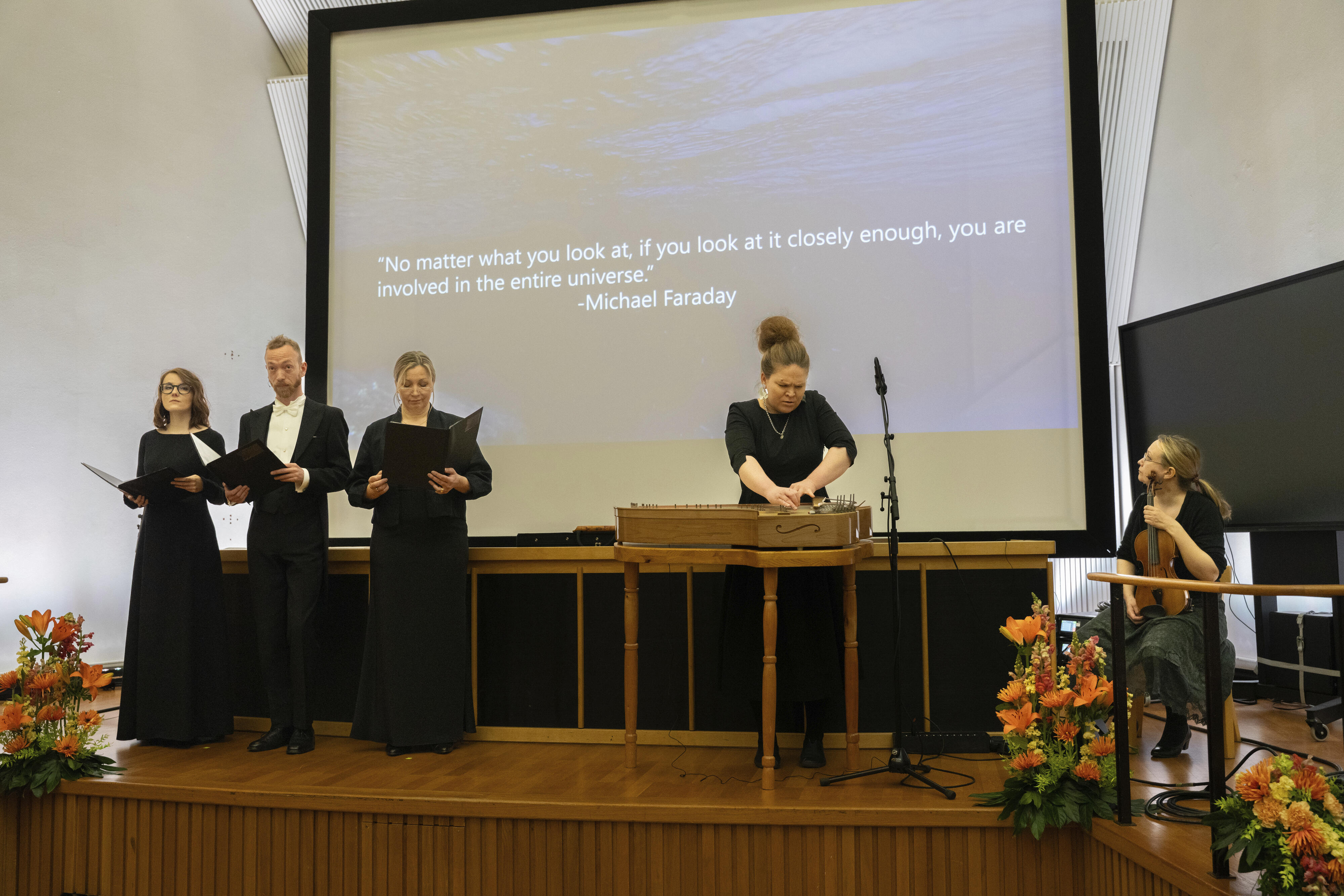 Group of people on a stage performing. Michael Faraday's quote is displayed behind them. Flower arrangements in front.