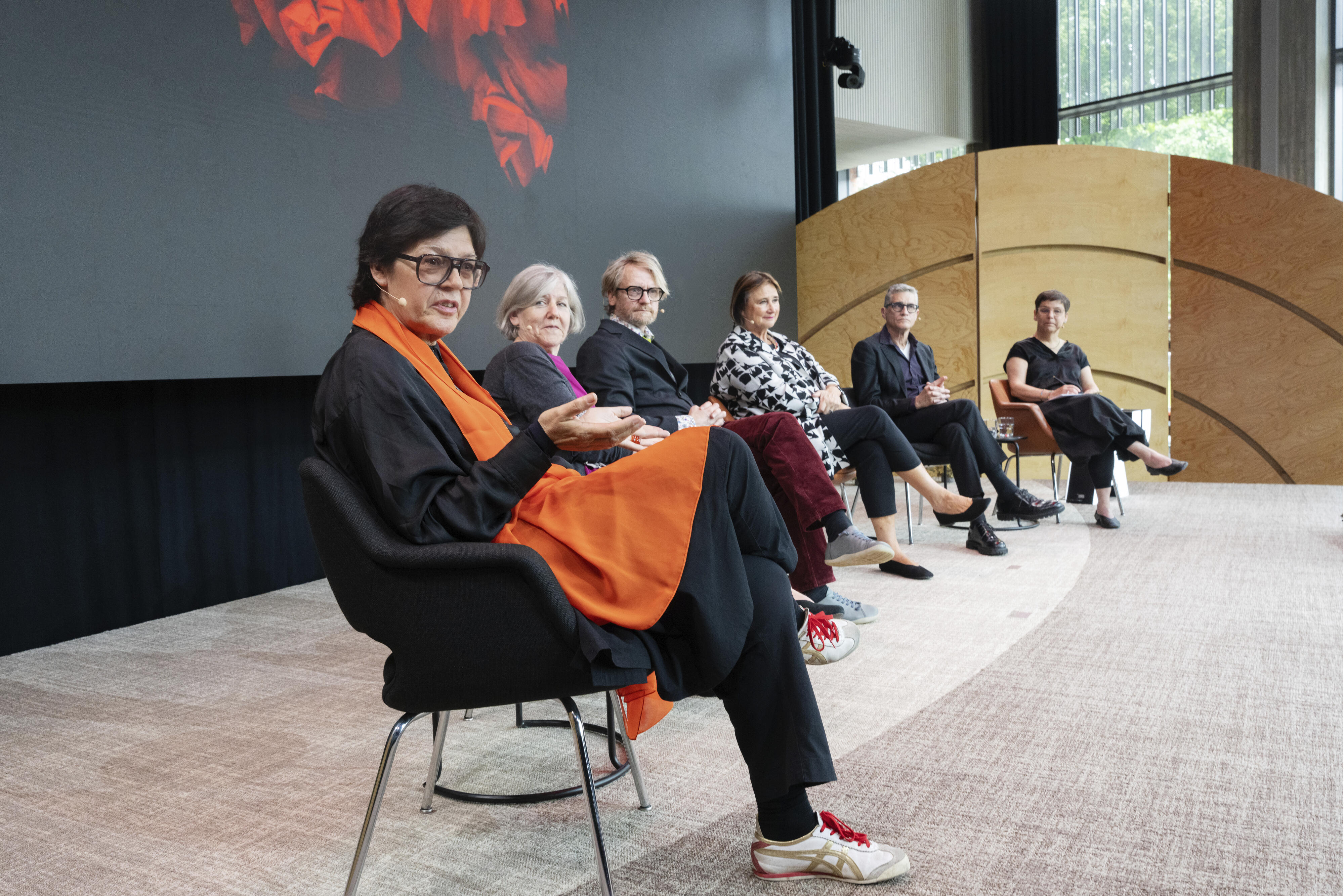 Kuusi ihmistä istuu paneelikeskustelussa. Etualalla oleva henkilö pukeutunut mustiin vaatteisiin ja oranssiin huiviin.